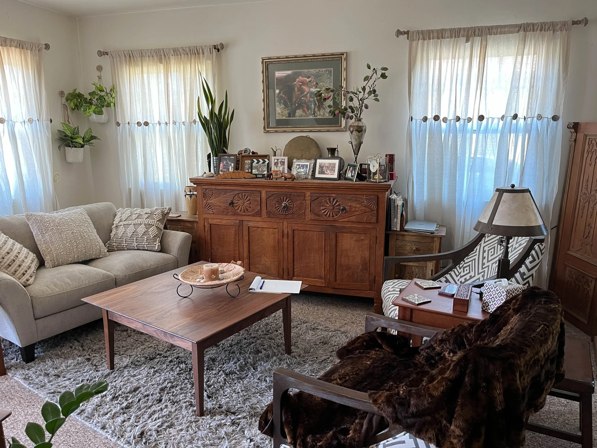 Living room with a beige sofa, a wooden coffee table with a candle, several framed photographs, and a decorative tray. A shaggy rug covers the floor. A wooden cabinet with decorative carvings and a table lamp with a patterned shade are on the right. Multiple windows with sheer curtains allow natural light. Inside the room, there are various potted plants and artwork on the wall.