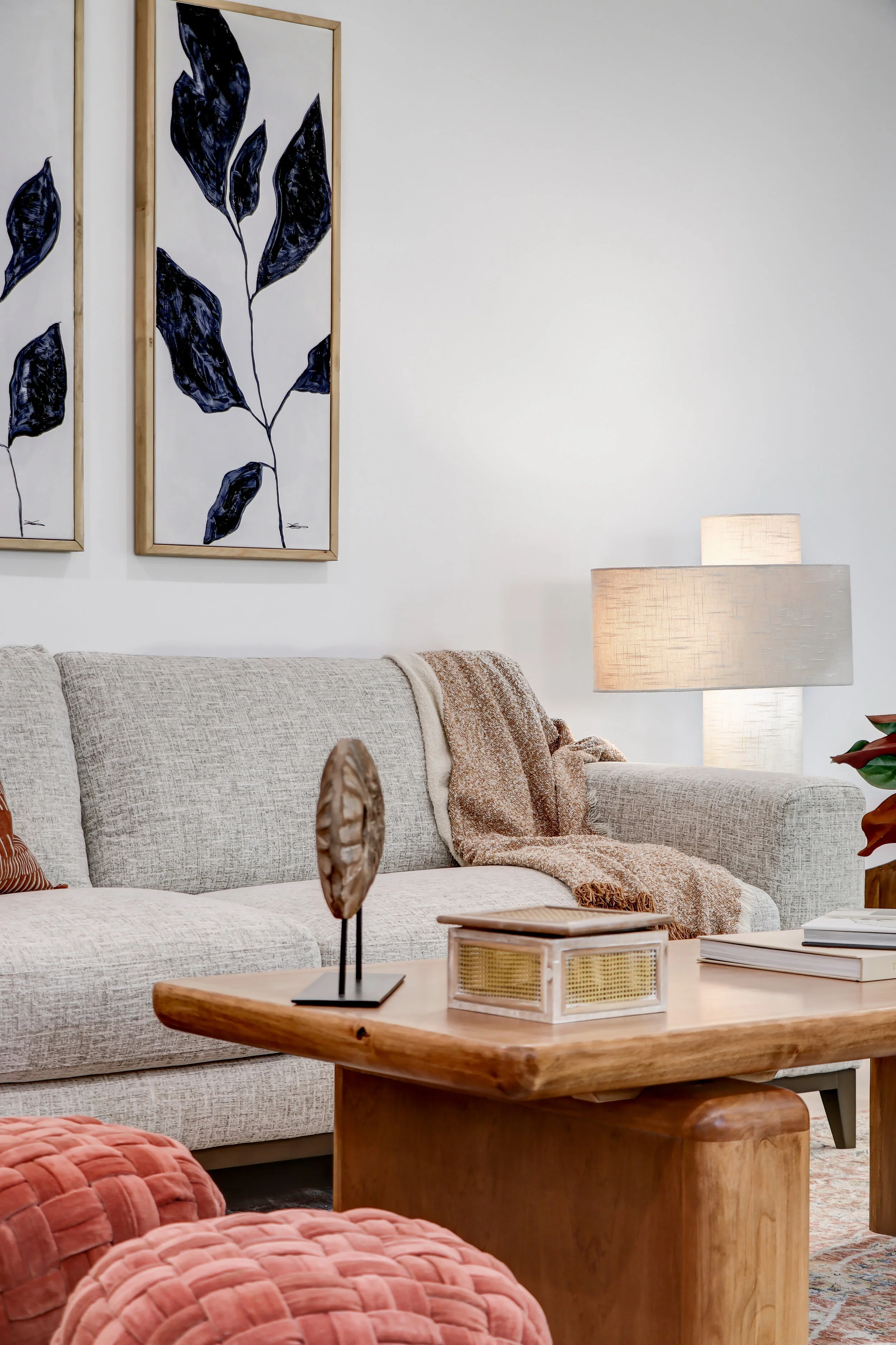 Living room with beige couch, a wooden coffee table, decorative objects, beige and pink pillows, a lamp, and wall art with dark leaf patterns.