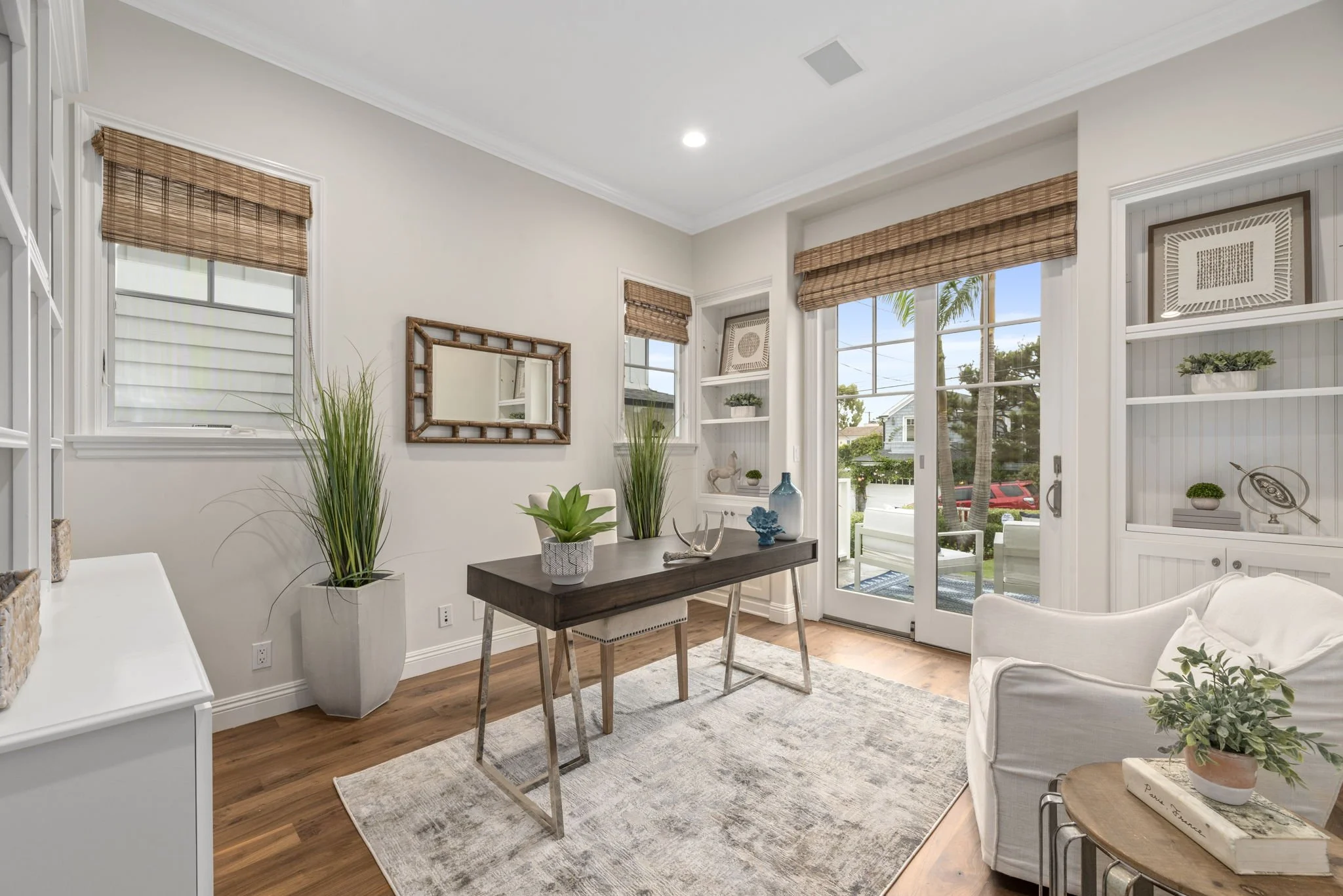 Bright living room with white walls, hardwood floors, and large windows with woven shades, decorated with houseplants, a mirror, and coastal-themed decor.