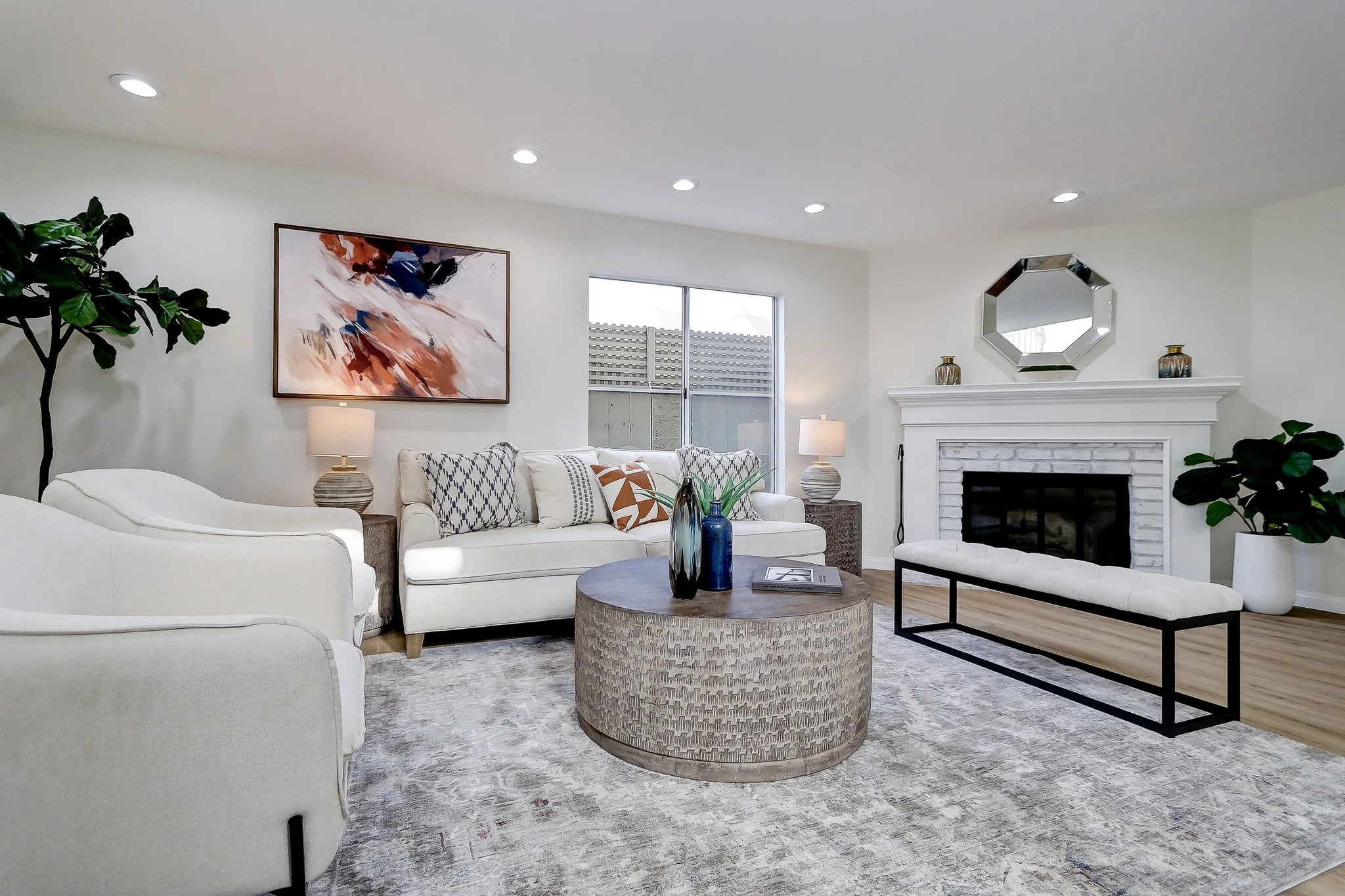 A modern living room with white couches, a round coffee table with blue vases, a fireplace with a mirror above, green plants, abstract wall art, lamps, and a window.