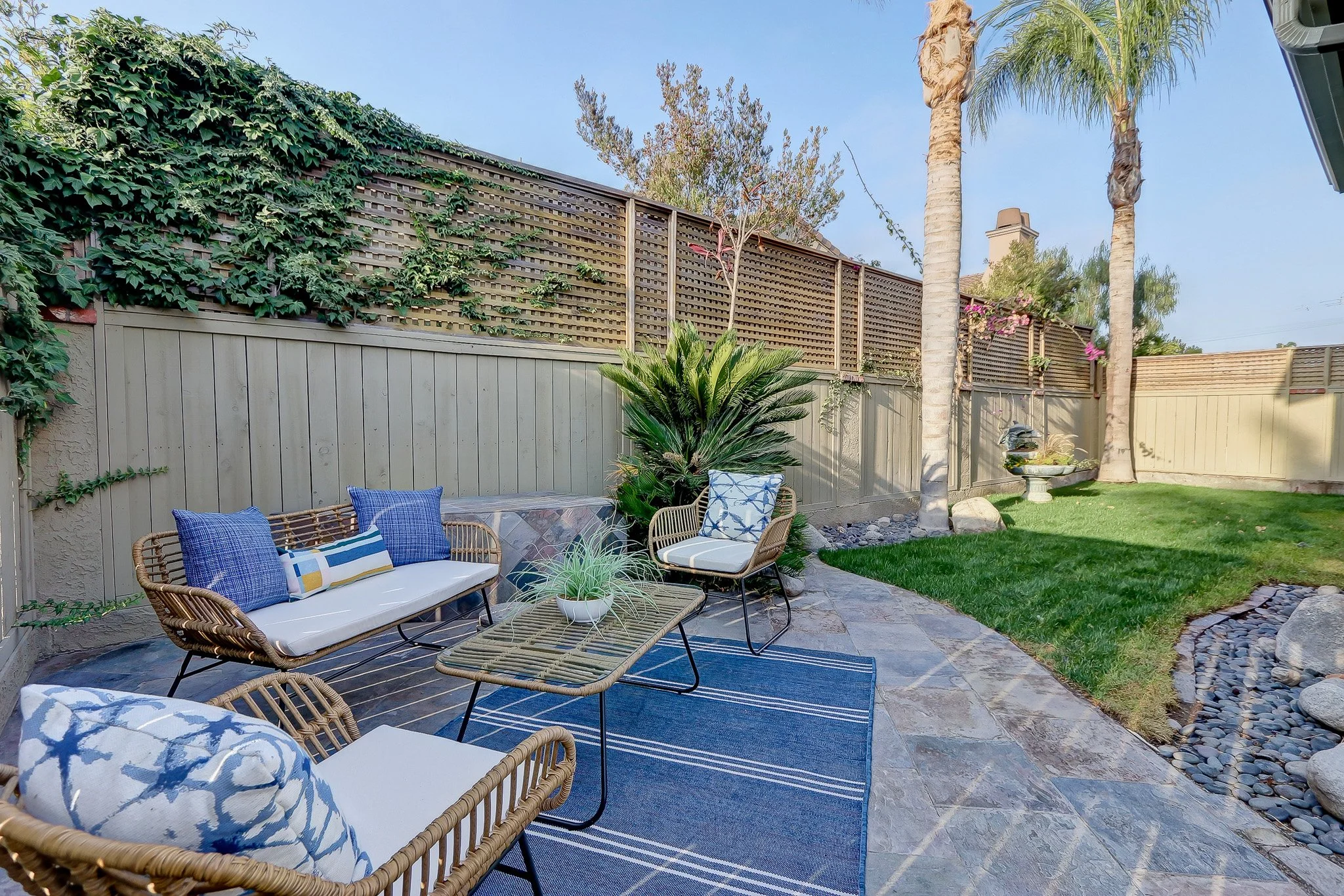 A backyard patio with wicker chairs and a bench, decorated with blue and patterned cushions, a small table with a potted plant, surrounded by a beige wooden fence, green plants, trees, and a lawn under a clear blue sky.