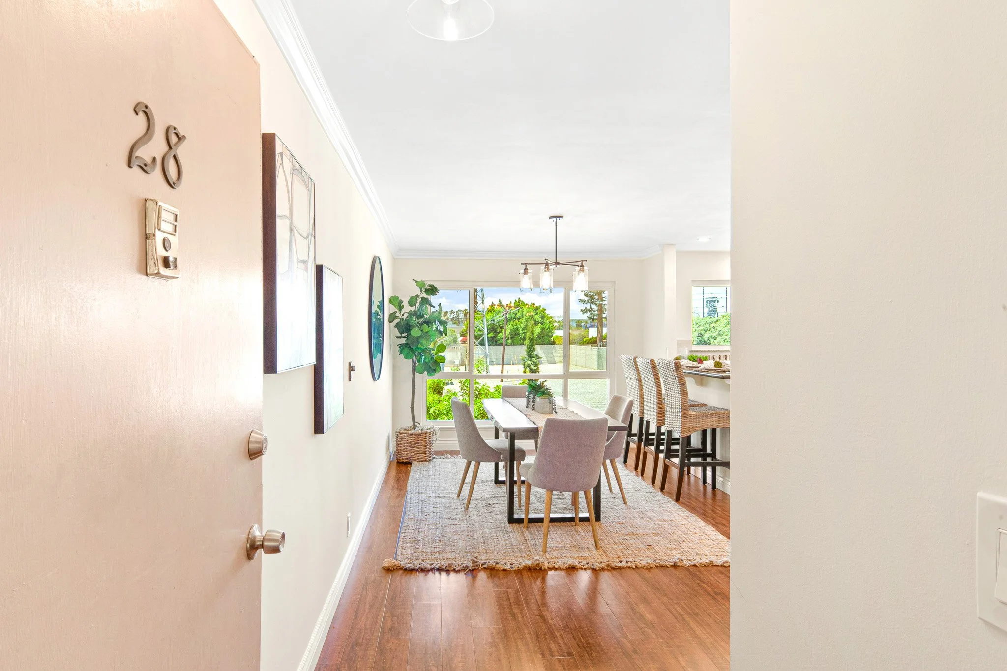 View of a dining and living area through the doorway, featuring a dining table with six chairs, a large window showing greenery outside, potted plants, and wall art.