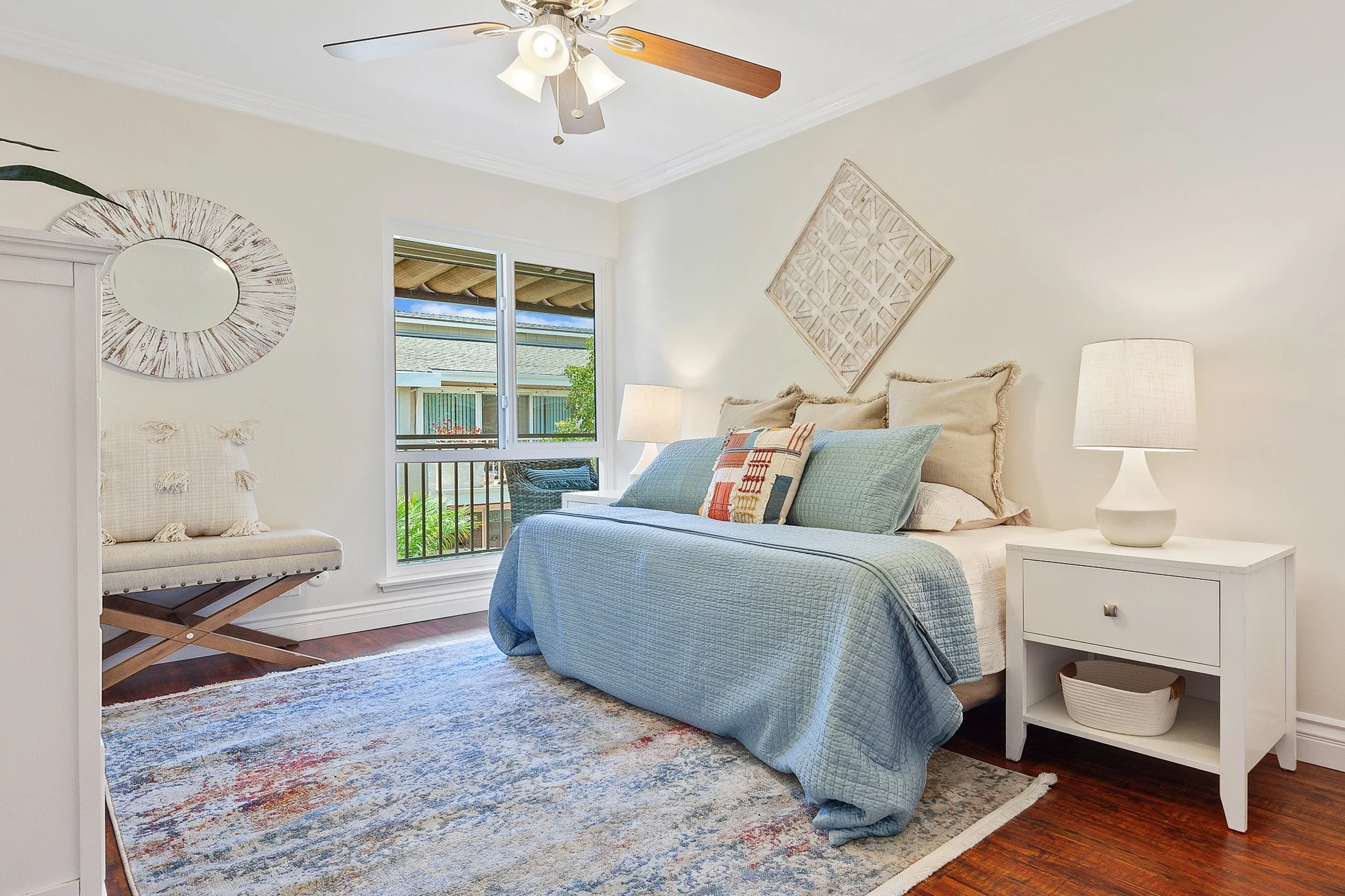 A bedroom with a bed, nightstand, lamps, wall decor, and a window showing the outside. The room has a ceiling fan and a colorful area rug.