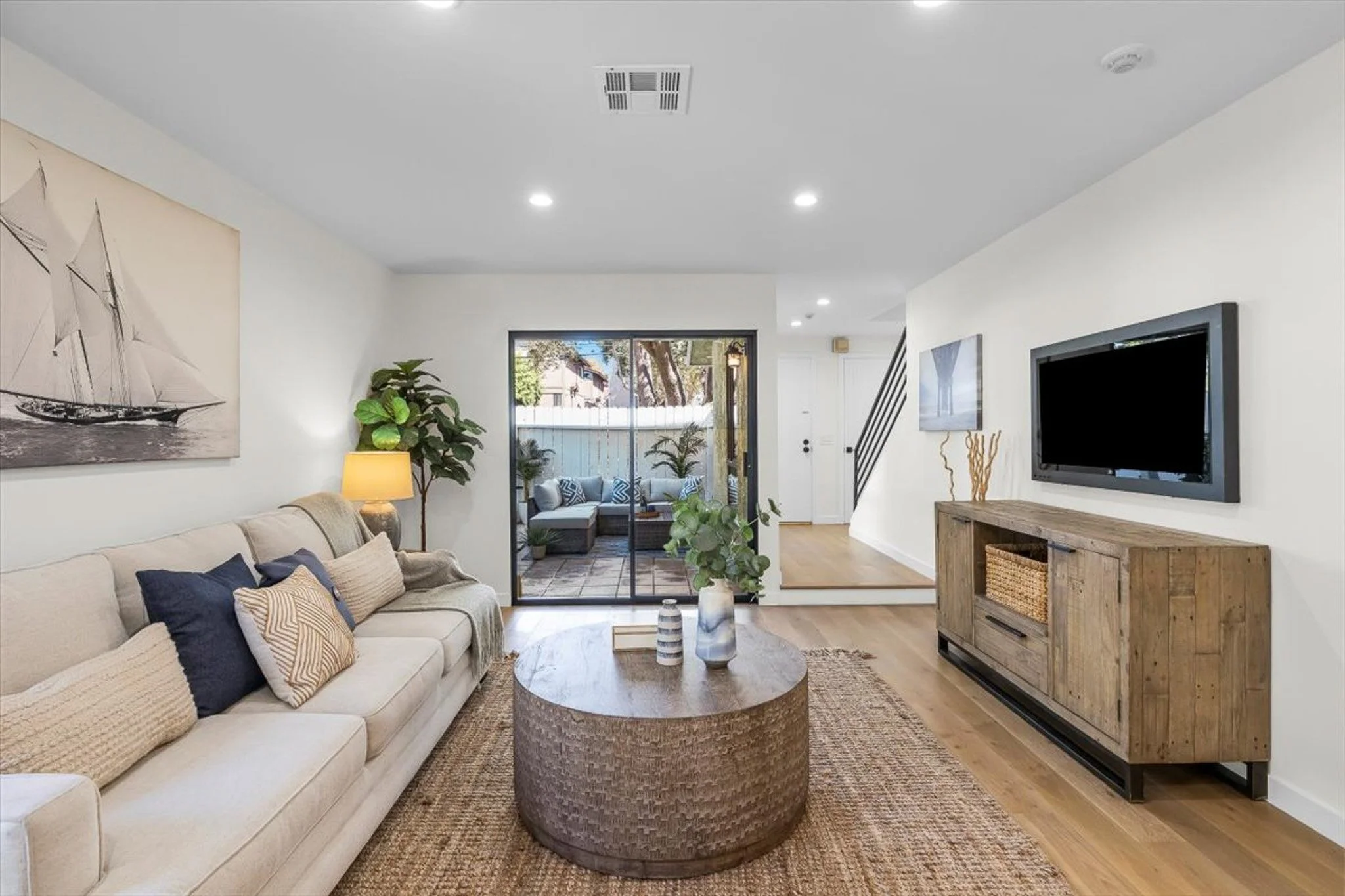 Living room with beige sofa, pillows, a wooden coffee table, a television mounted on the wall, potted plants, and a view of a patio with outdoor furniture through sliding glass doors.