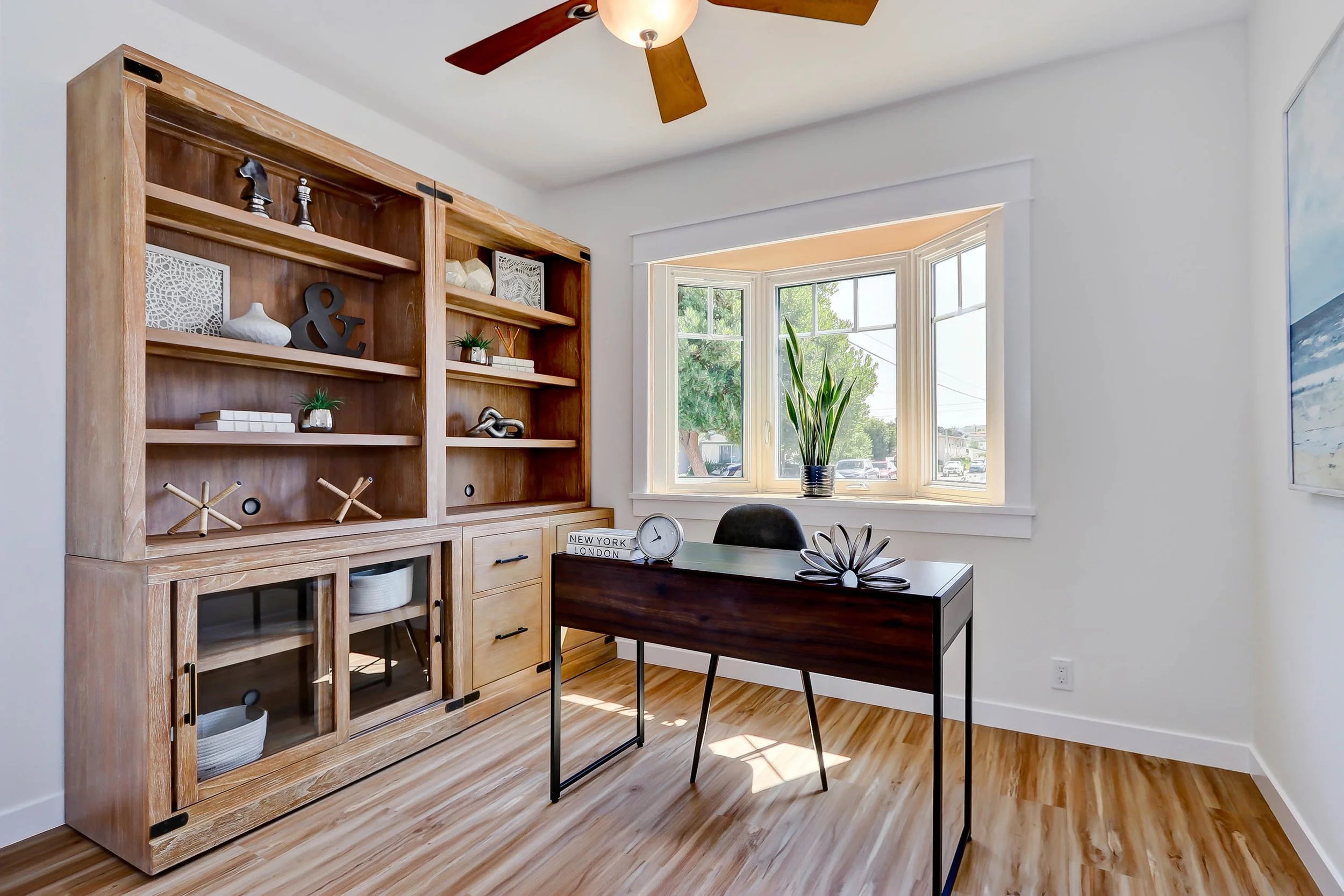 A room with a wooden bookshelf, a table, chairs, a large window with a plant on the windowsill, and a ceiling fan.