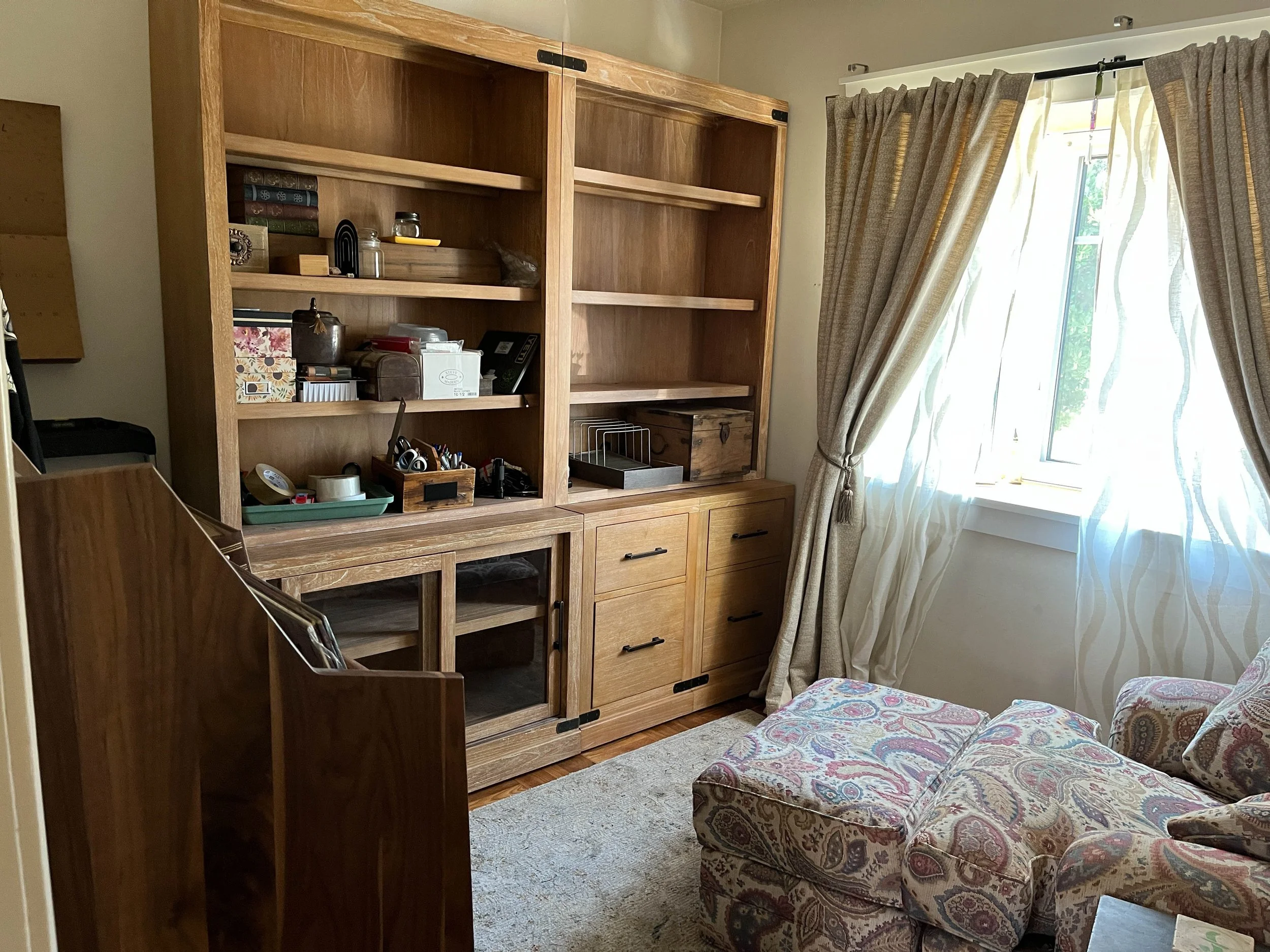 A wooden bookcase with sliding doors, partially open, displaying various items like books, jars, boxes, and office supplies. Next to the bookcase is a window with beige curtains, allowing sunlight to brighten the room. There is a patterned armchair with floral upholstery and an ottoman visible in the lower right corner, and a carpet on the floor.