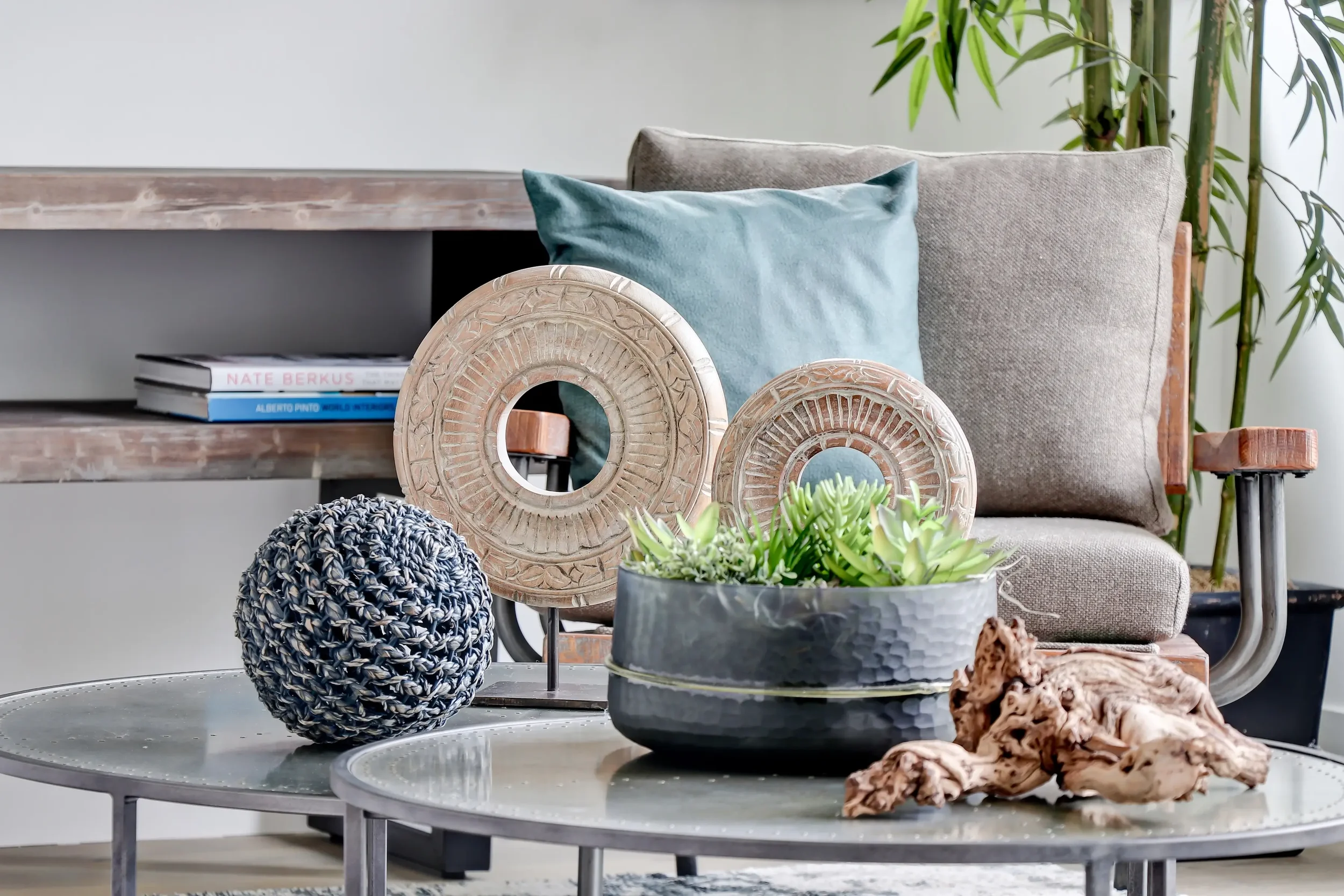 Decorative objects on a gray table, including two carved circular mirrors, a knitted black and white ball, a black and gray planter with green succulent plants, and pieces of natural driftwood. In the background, a beige fabric armchair with a light blue pillow, a wooden side table, a shelf with books, and a tall plant with green leaves.