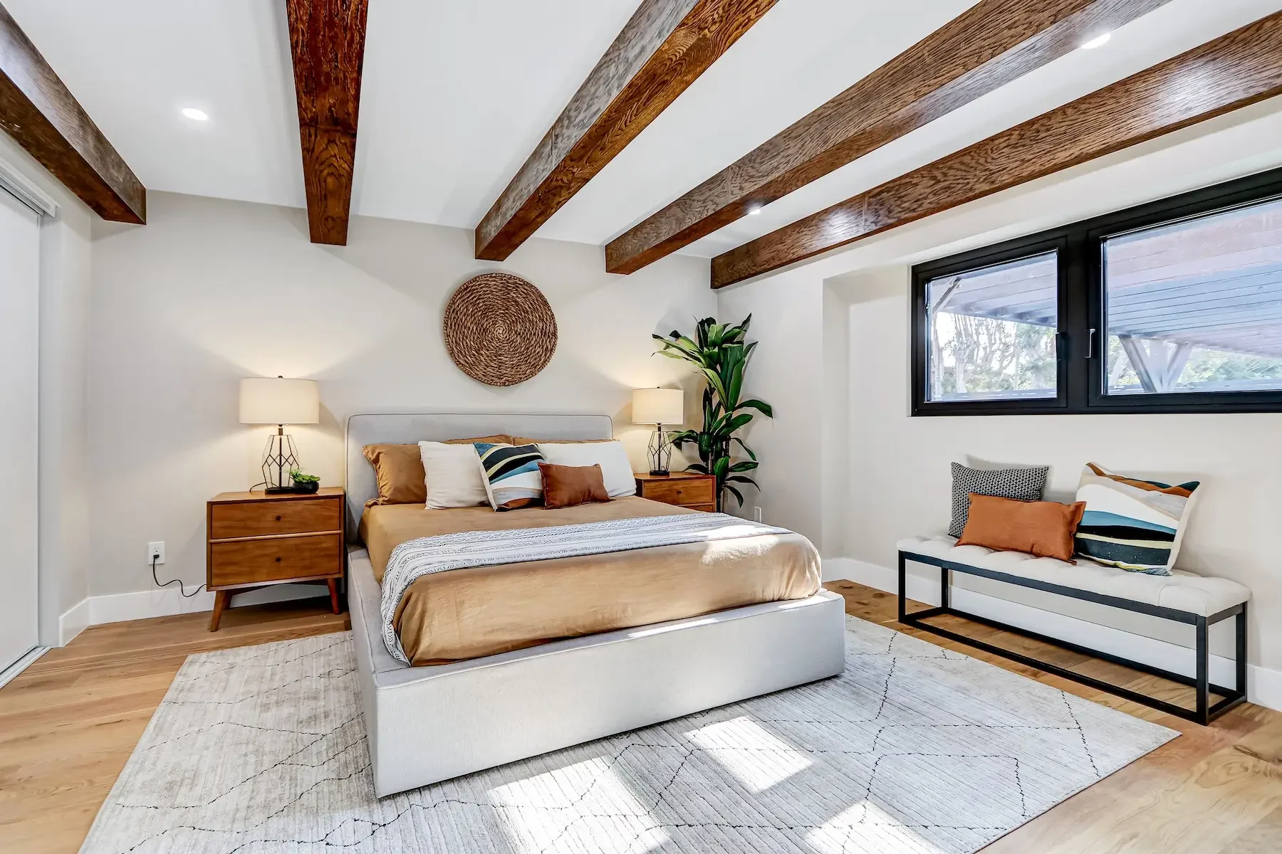 A modern bedroom with a white bed, wooden nightstands, lamps, a large window, a white bench with cushions, a green plant, and wooden ceiling beams.
