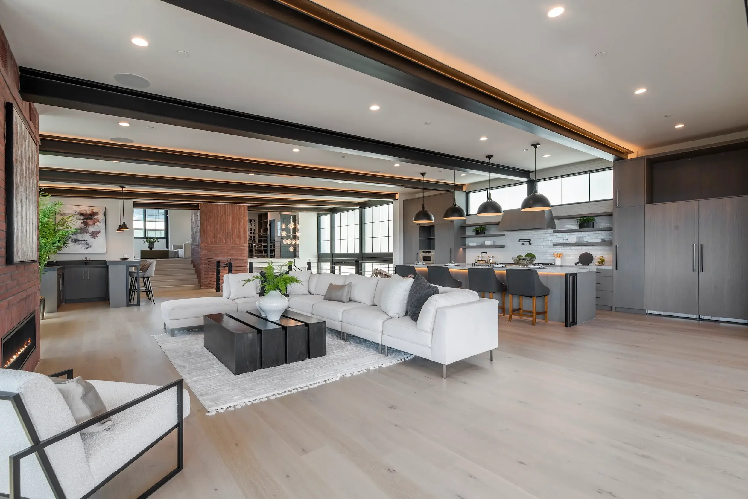 Modern open-concept living room and kitchen with white sofa, black coffee table, light wood flooring, gray cabinets, black pendant lights, large windows, and minimal decor.