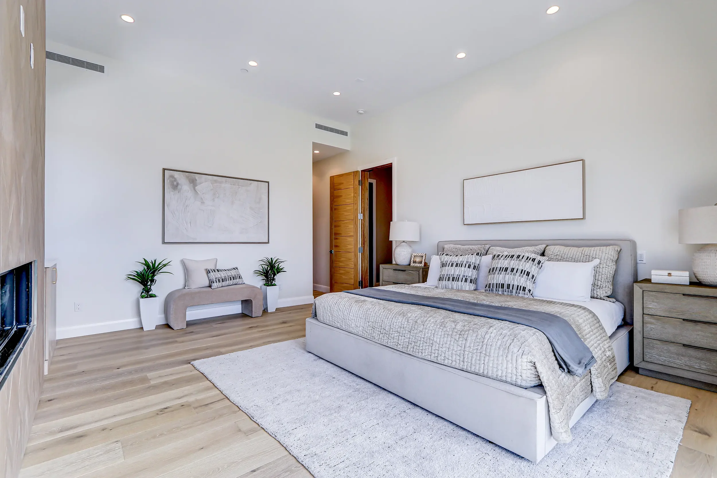 Modern bedroom with a large bed, light-colored bedding, side tables with lamps, abstract artwork, and small plants, with wood flooring and a partially open door.