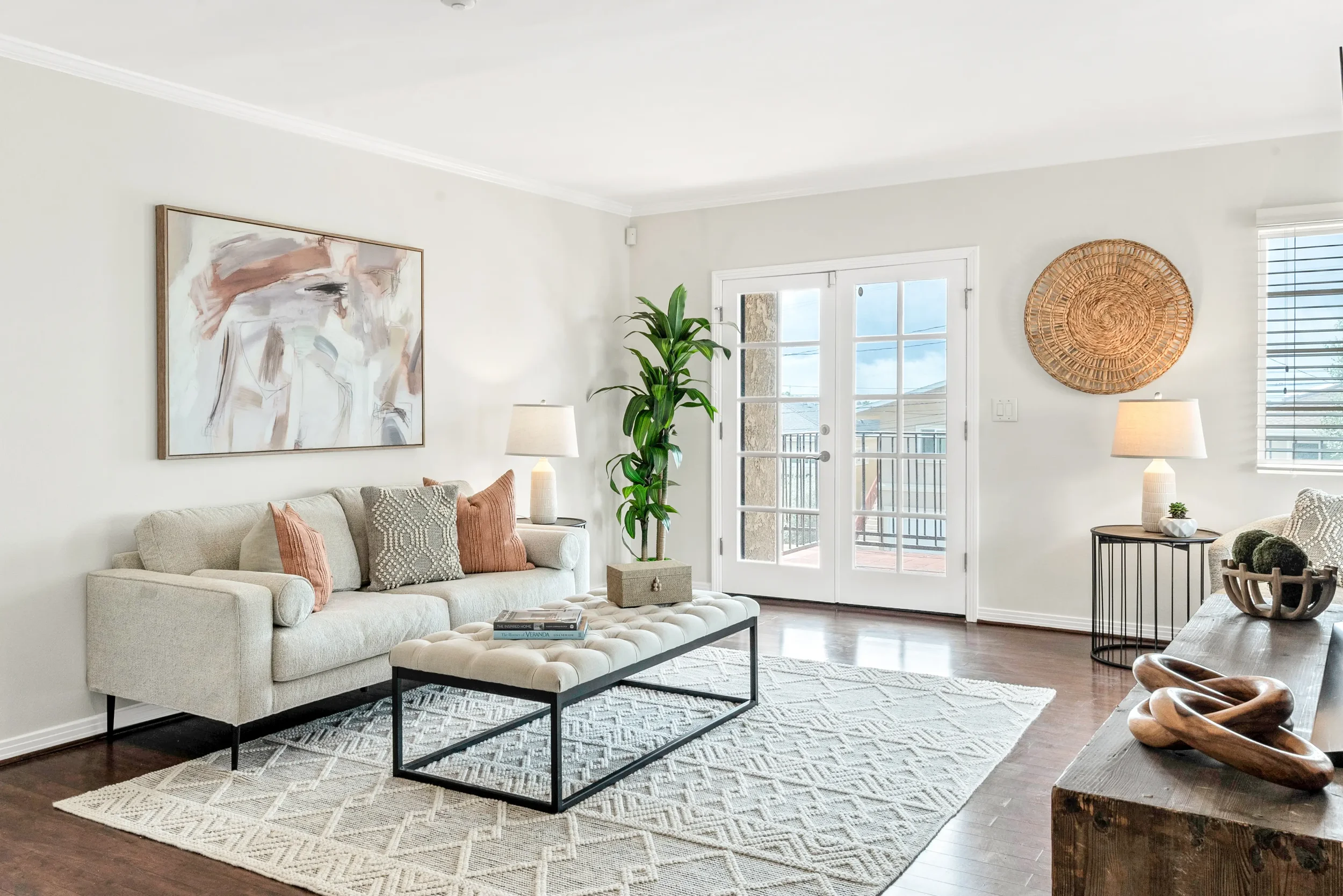 Living room with beige sofa, patterned pillows, abstract art on cream wall, tall green plant, glass door with balcony, two table lamps, woven wall decor, window blinds, wooden coffee table with sculptures, patterned rug, and hardwood floor.