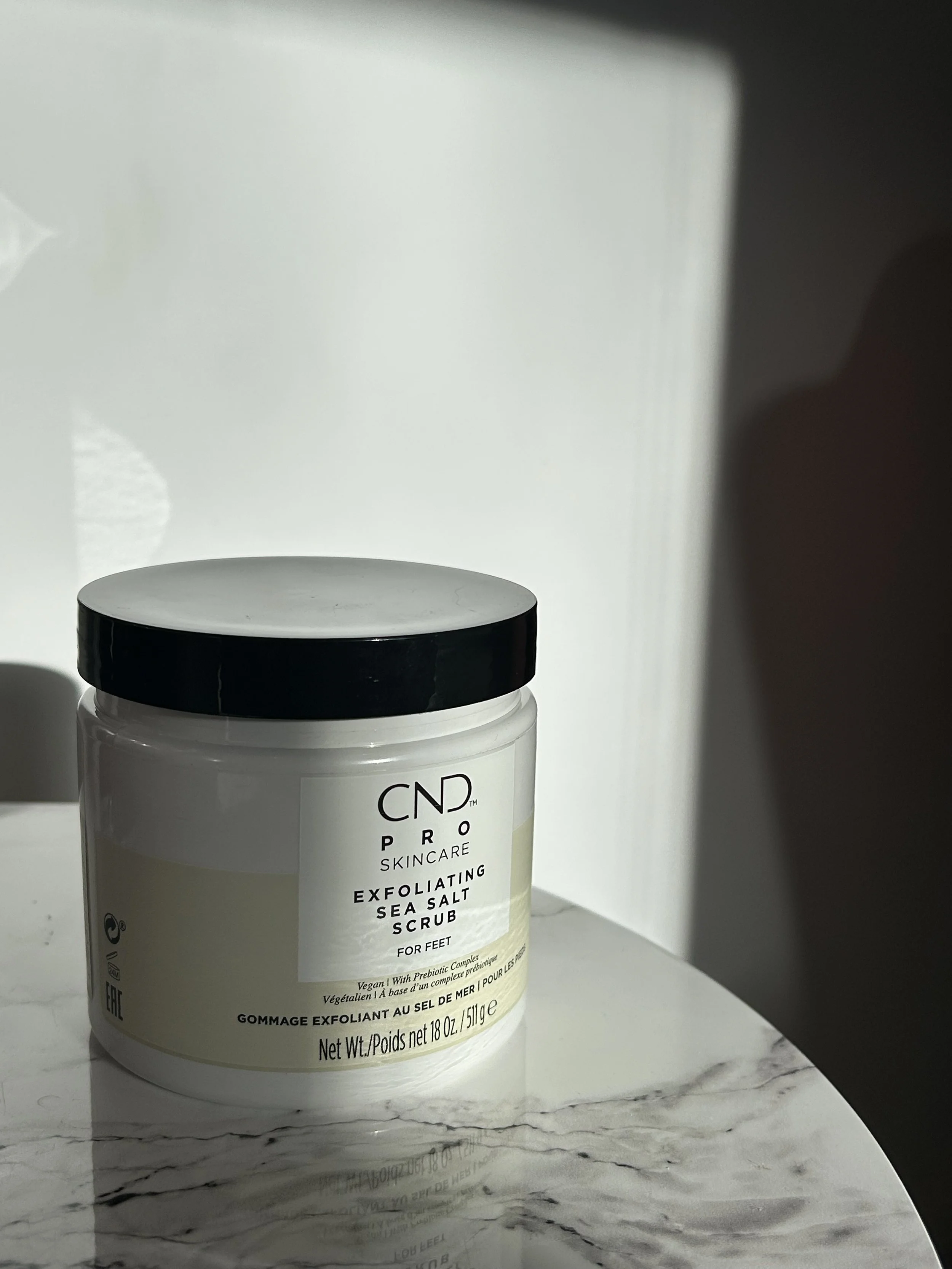 A container of CND Pro Skincare exfoliating sea salt scrub with a black lid, placed on a white marble surface with a white background.