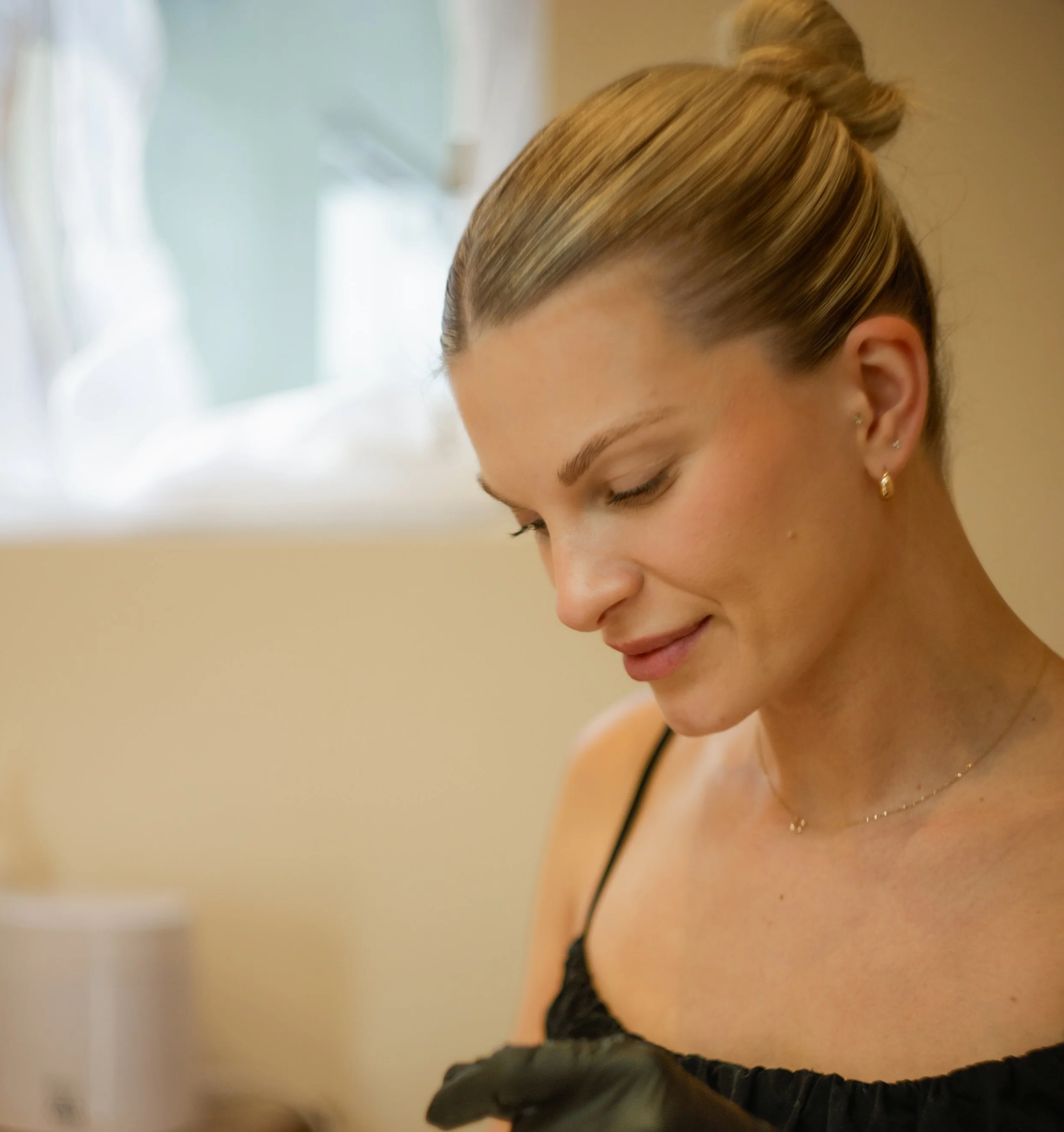 Close-up of a woman with blonde hair tied in a bun, wearing earrings, with a black strap top, smiling gently, with a soft background.