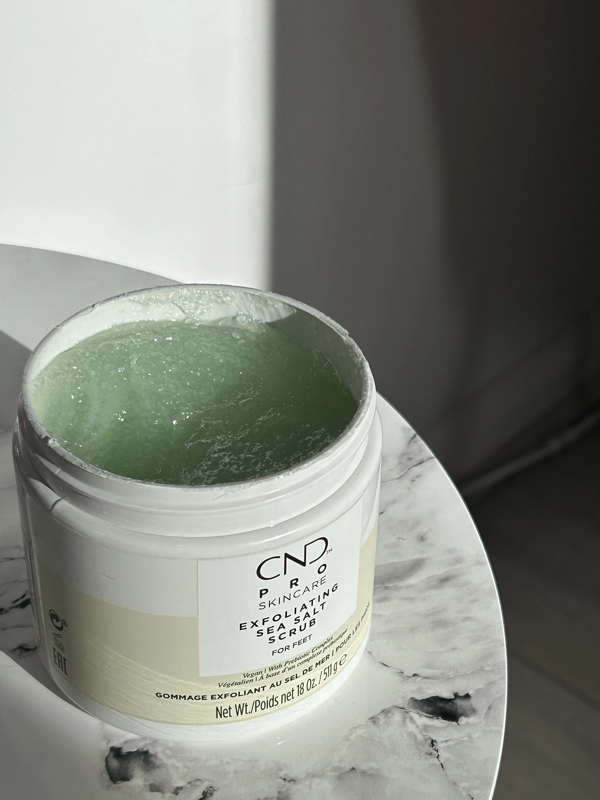 Container of exfoliating sea salt scrub on a marble surface with white background.
