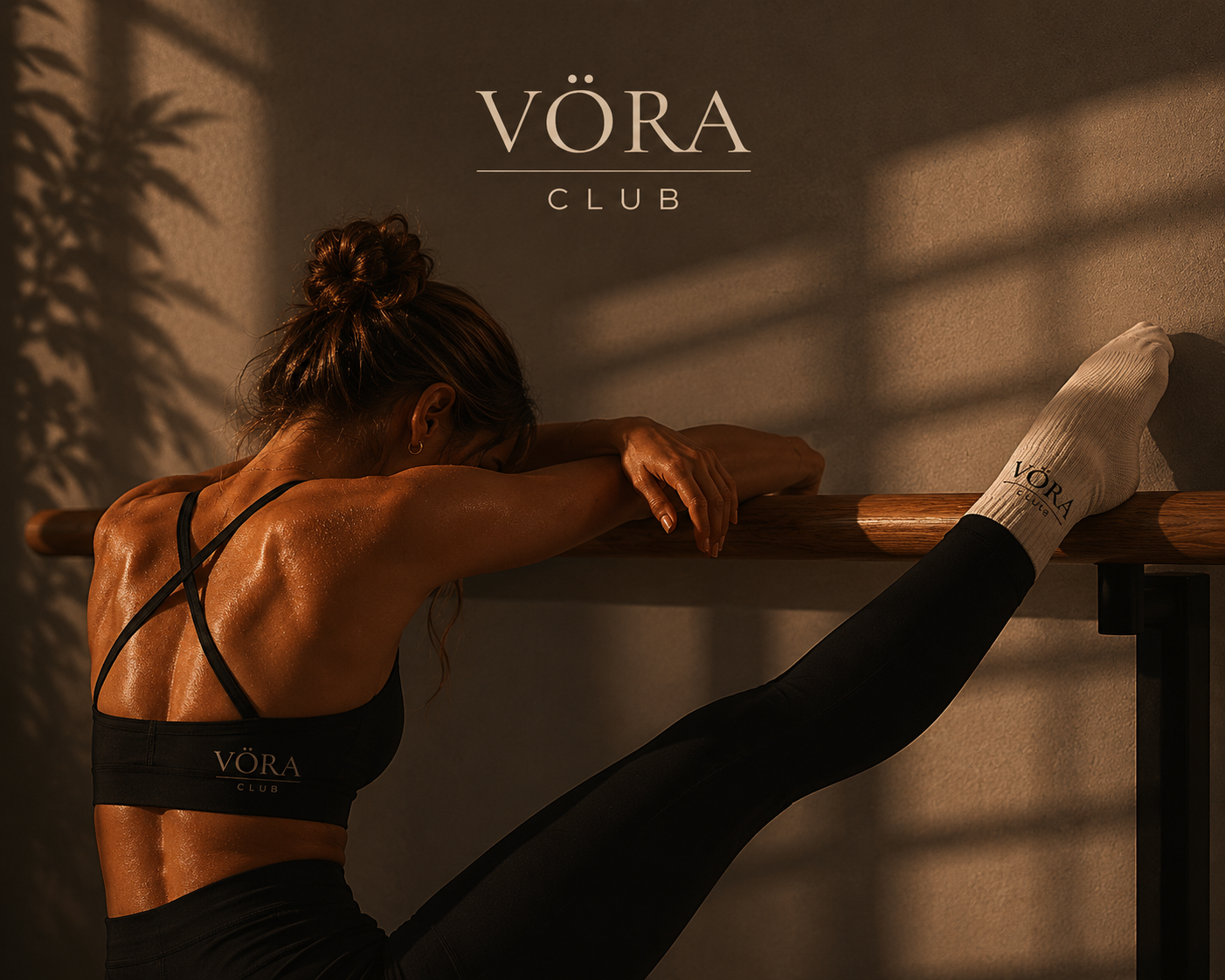 Una mujer haciendo ejercicio en un gimnasio, apoyada en una barra de ballet, con un pantalón y top negros que llevan la marca VÖRA CLUB, en un ambiente con luz cálida y sombras.