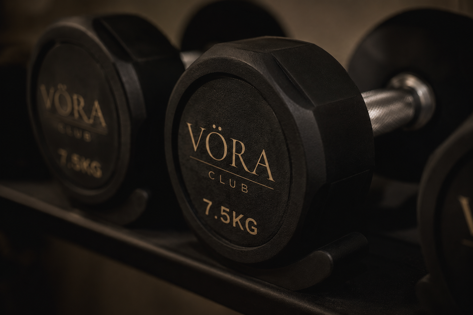 Mancuernas negras con peso de 7.5kg y la marca VÖRA en un rack de pesas en un gimnasio.