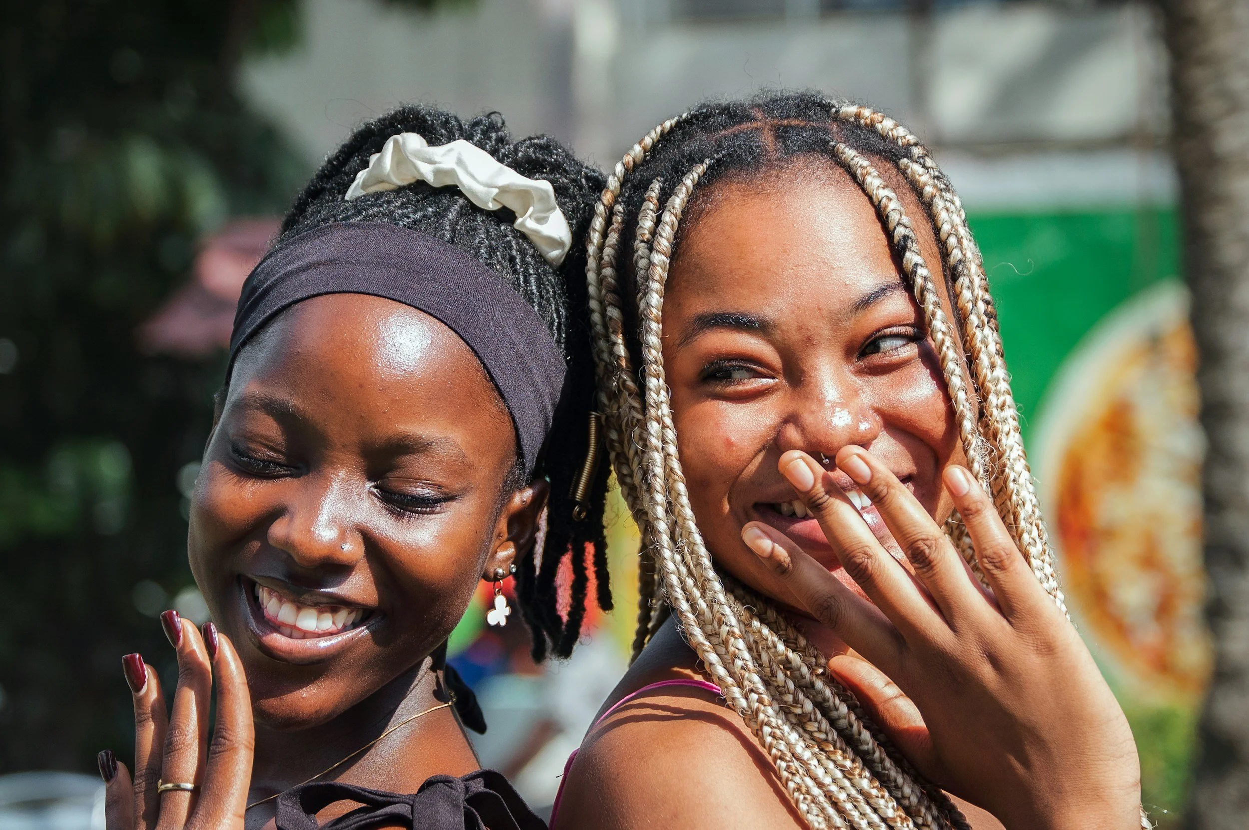 Two women smiling and laughing outdoors, one with braided hair and the other with natural curly hair, both dressed casually.