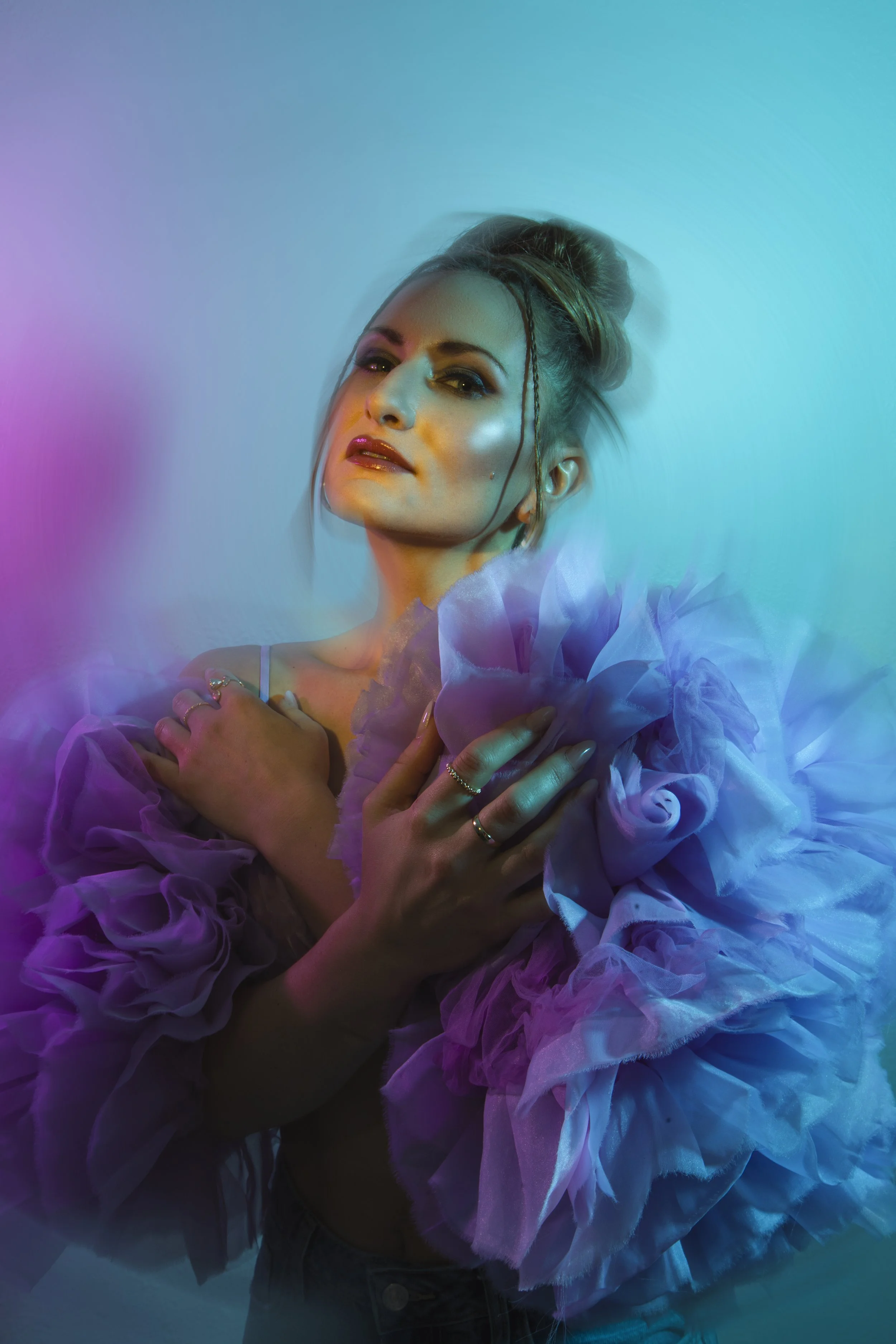 A woman with styled hair and makeup posing with her hands crossed over her chest, wearing a fluffy, ruffled garment, illuminated by colorful, vibrant lighting.