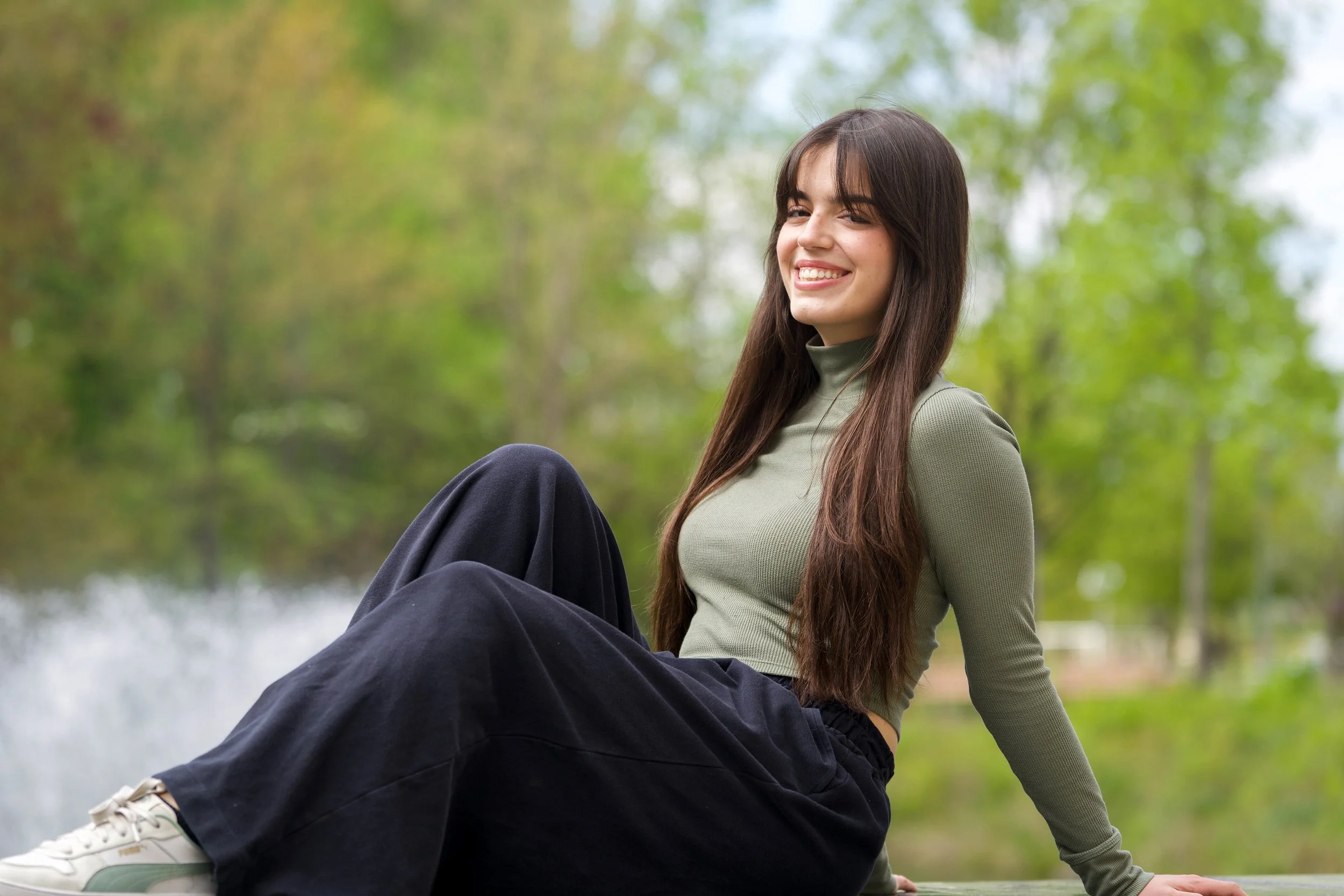 A young woman with long dark hair, smiling while sitting outdoors with green trees in the background. She is wearing a green long-sleeve shirt and black pants.