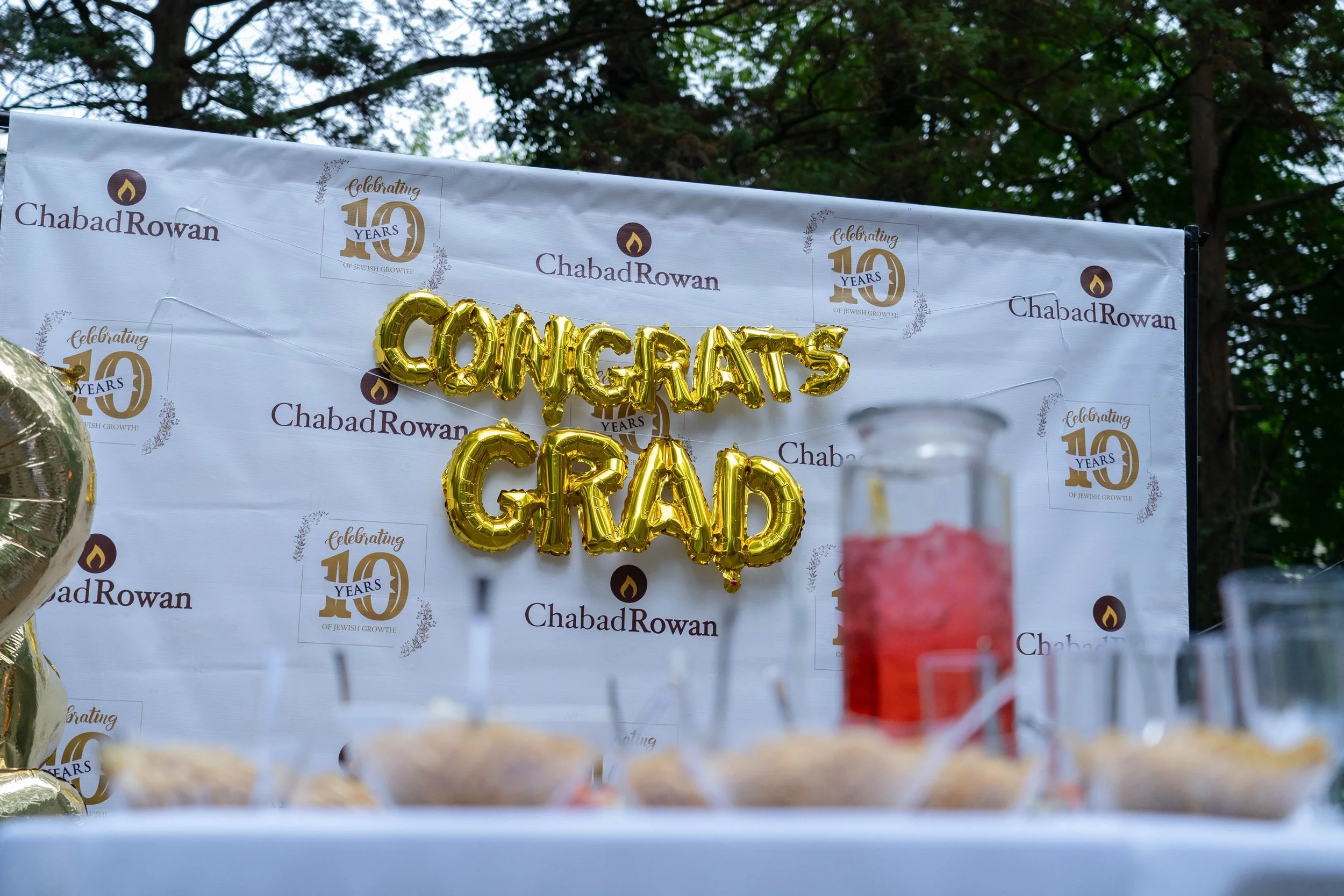 Celebration decorations with gold metallic balloons spelling 'CONGRATS' and a backdrop with Chabad Rowan logos and a '10 Years' logo, outdoors with trees in the background.