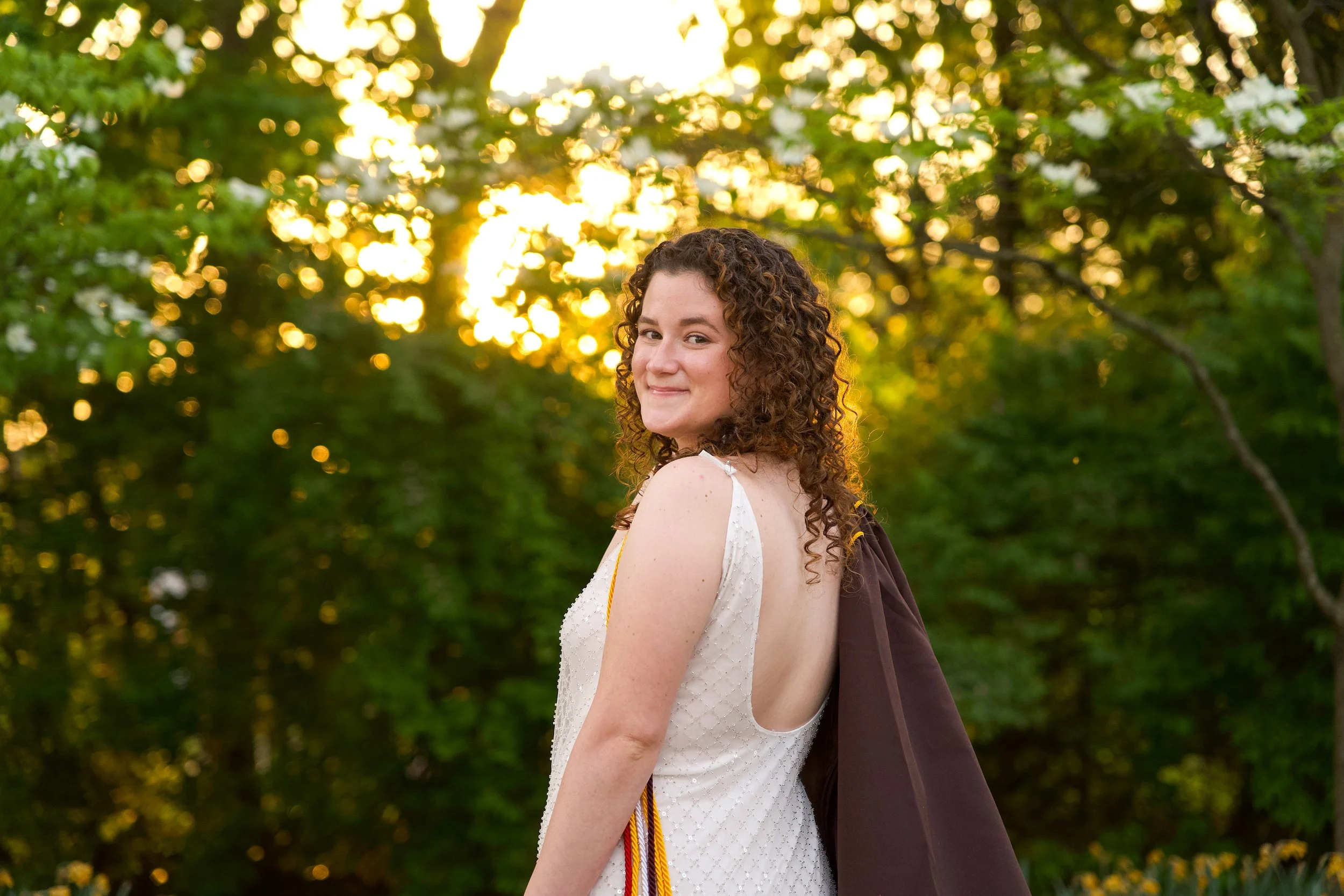 A young woman with curly hair in a white dress standing outdoors during sunset, smiling at the camera with green trees and sunlight in the background.