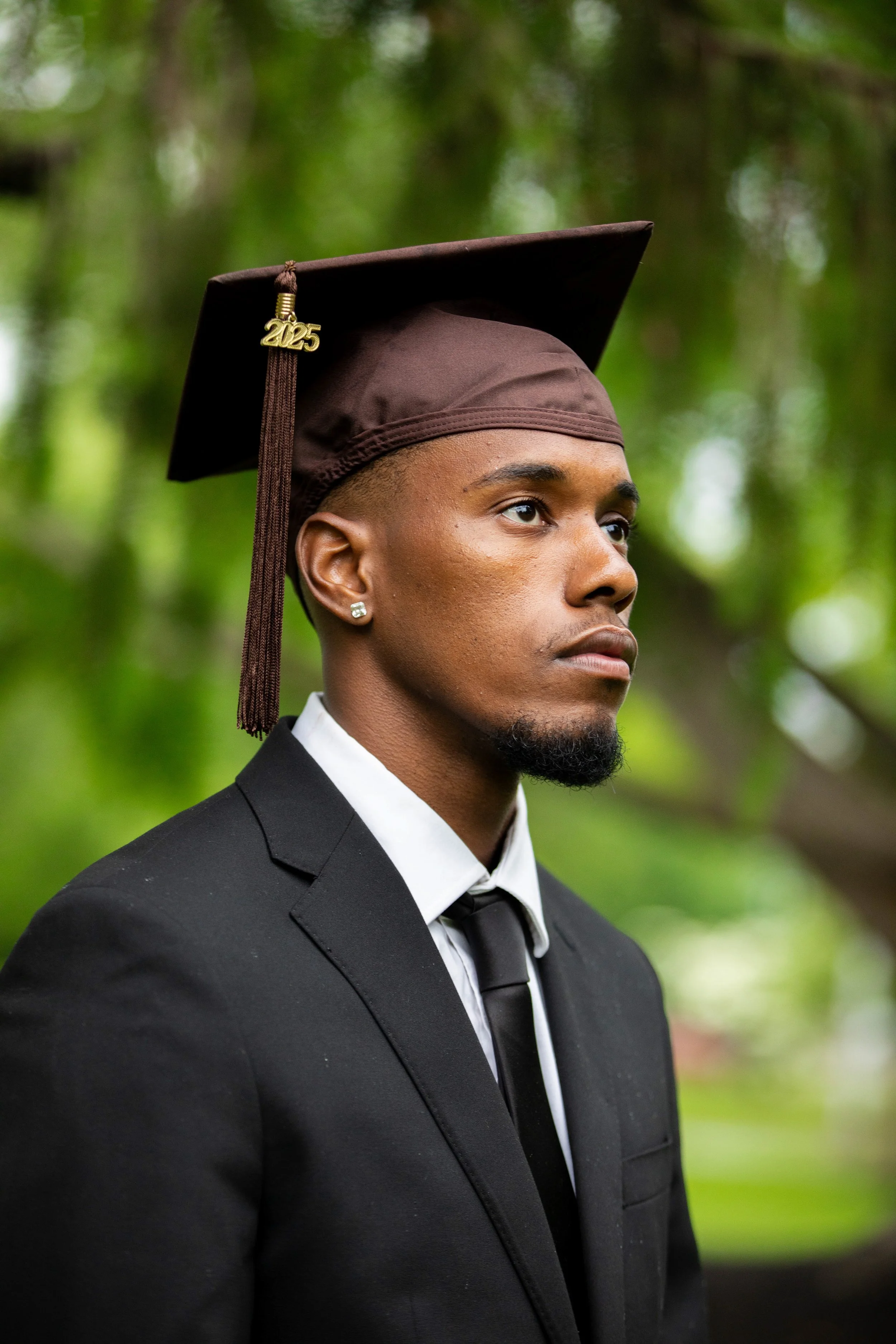 A young man in a black tuxedo and a white shirt, wearing a brown graduation cap with a "2025" tassel, standing outdoors with a blurred green background.