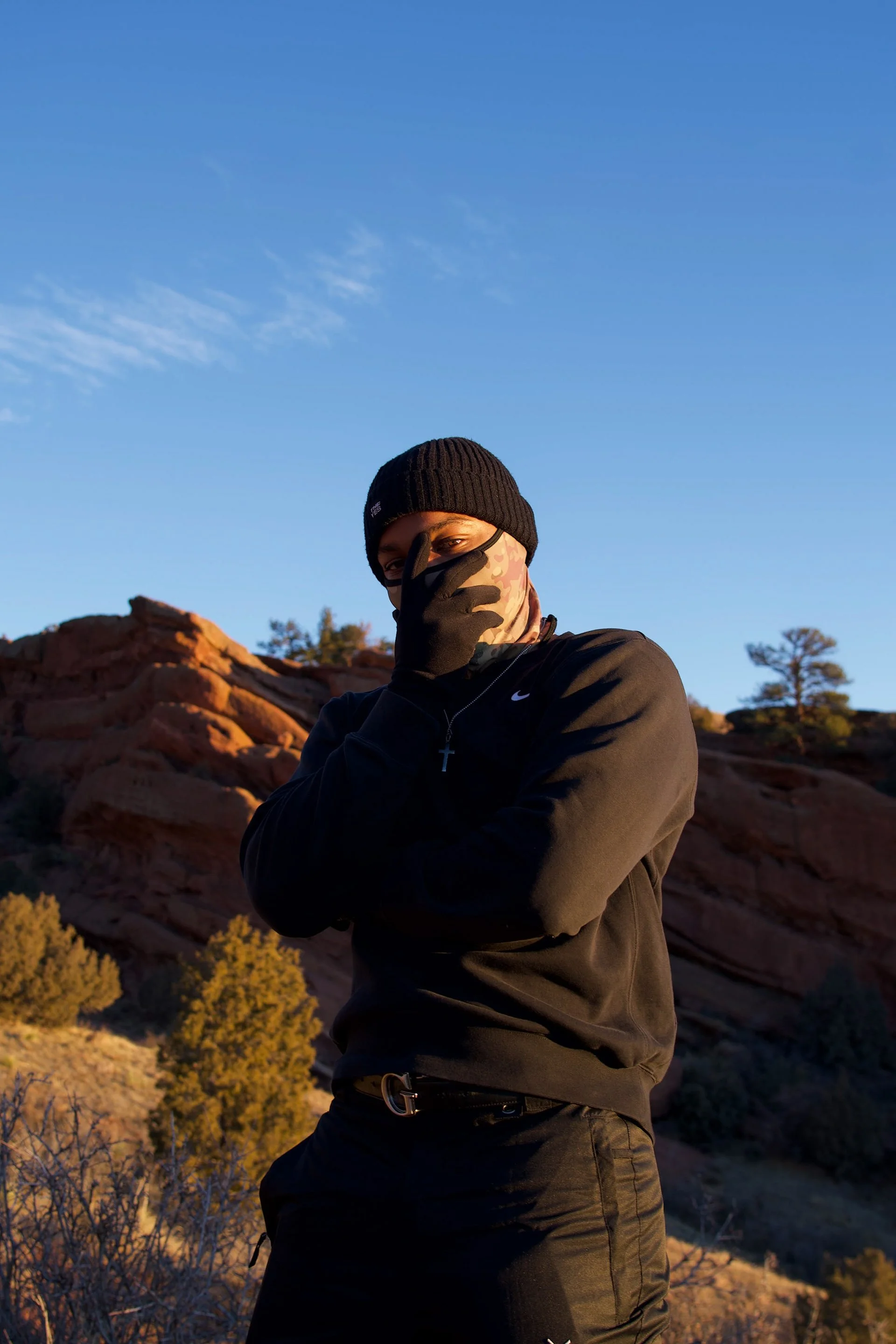 A person standing outdoors in front of a rocky landscape with trees, wearing a black beanie, black face mask, black jacket, and black pants, with a blue sky and some clouds overhead.