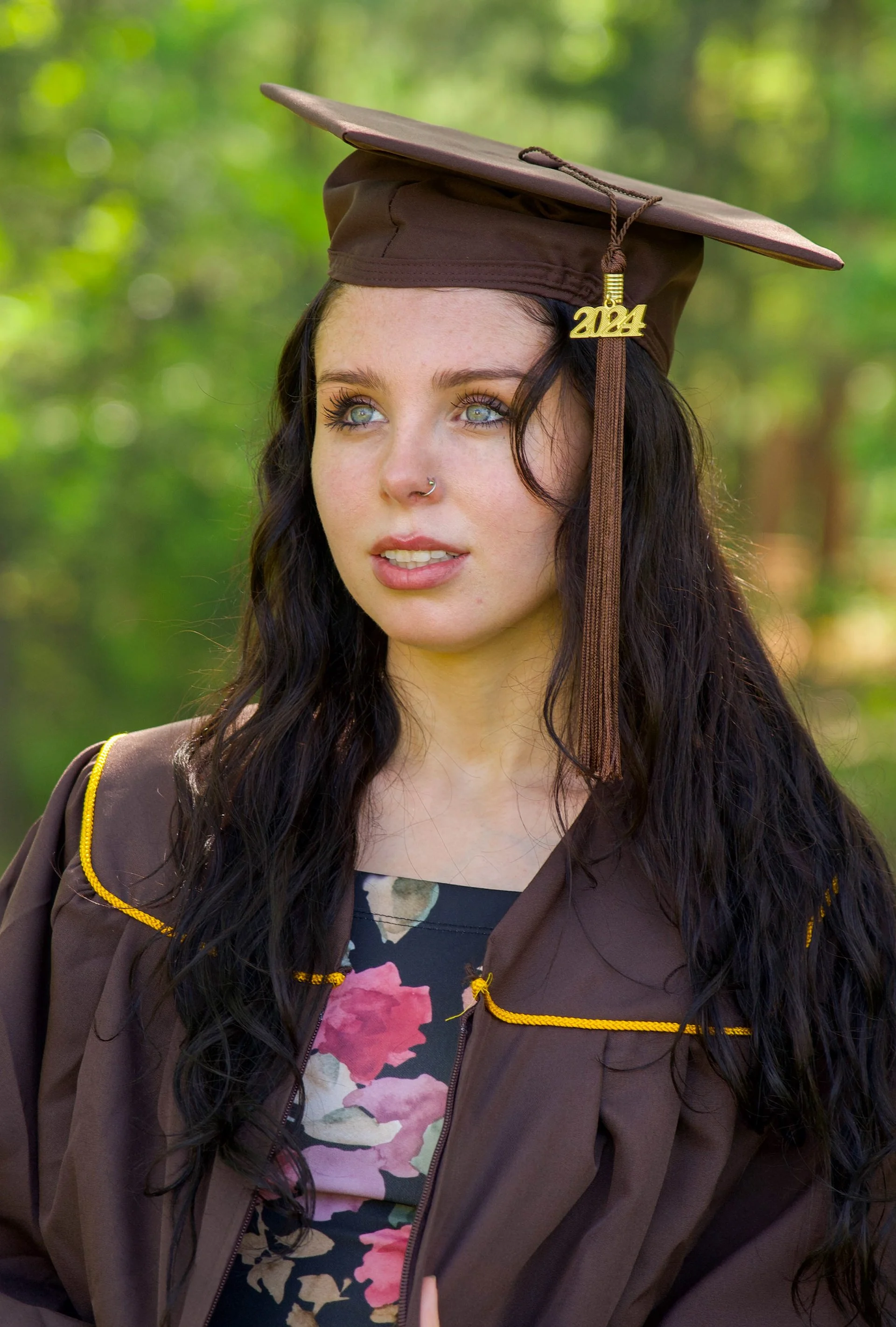 A young woman in a graduation cap and gown standing outdoors with a blurred green background.