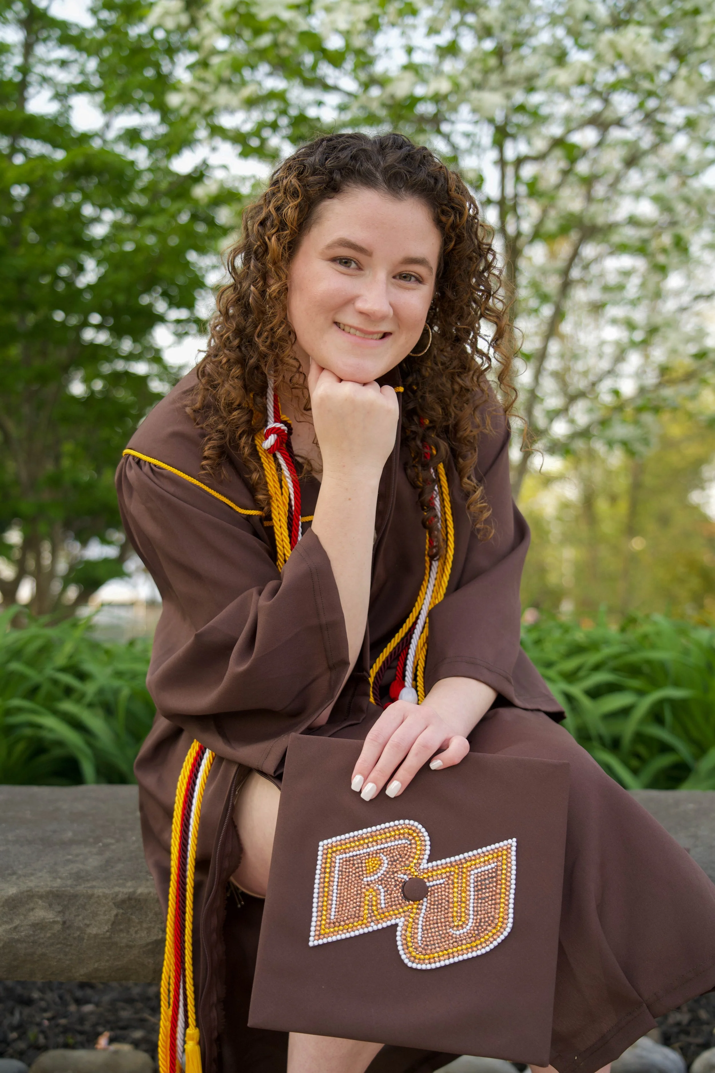 A young woman with curly brown hair smiling outdoors in a graduation gown and cap, holding a decorated diploma cover, with trees and greenery in the background.