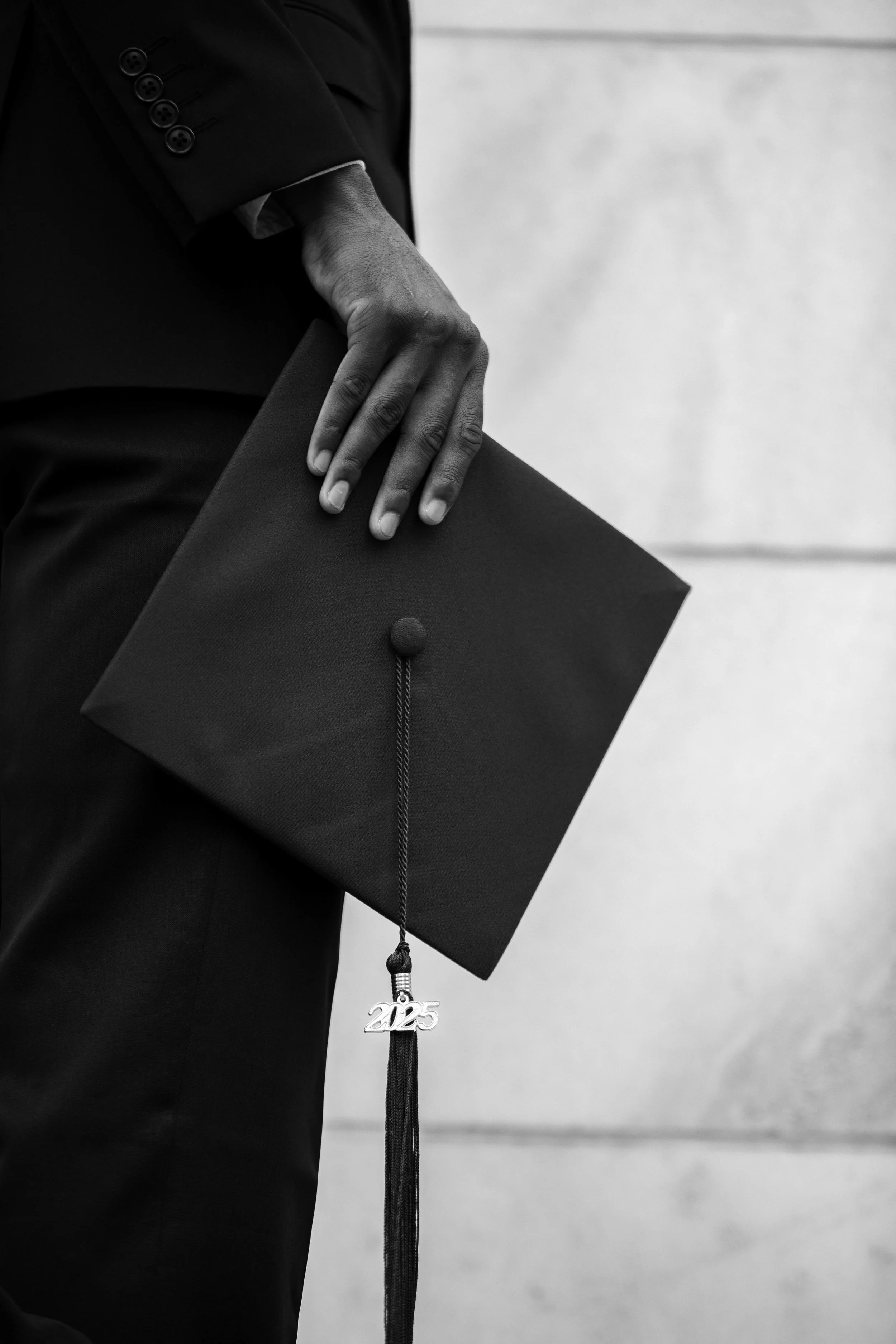 Close-up of a person in a suit holding a graduation cap with a "2025" tassel. The person appears to be sitting, with a plain background.