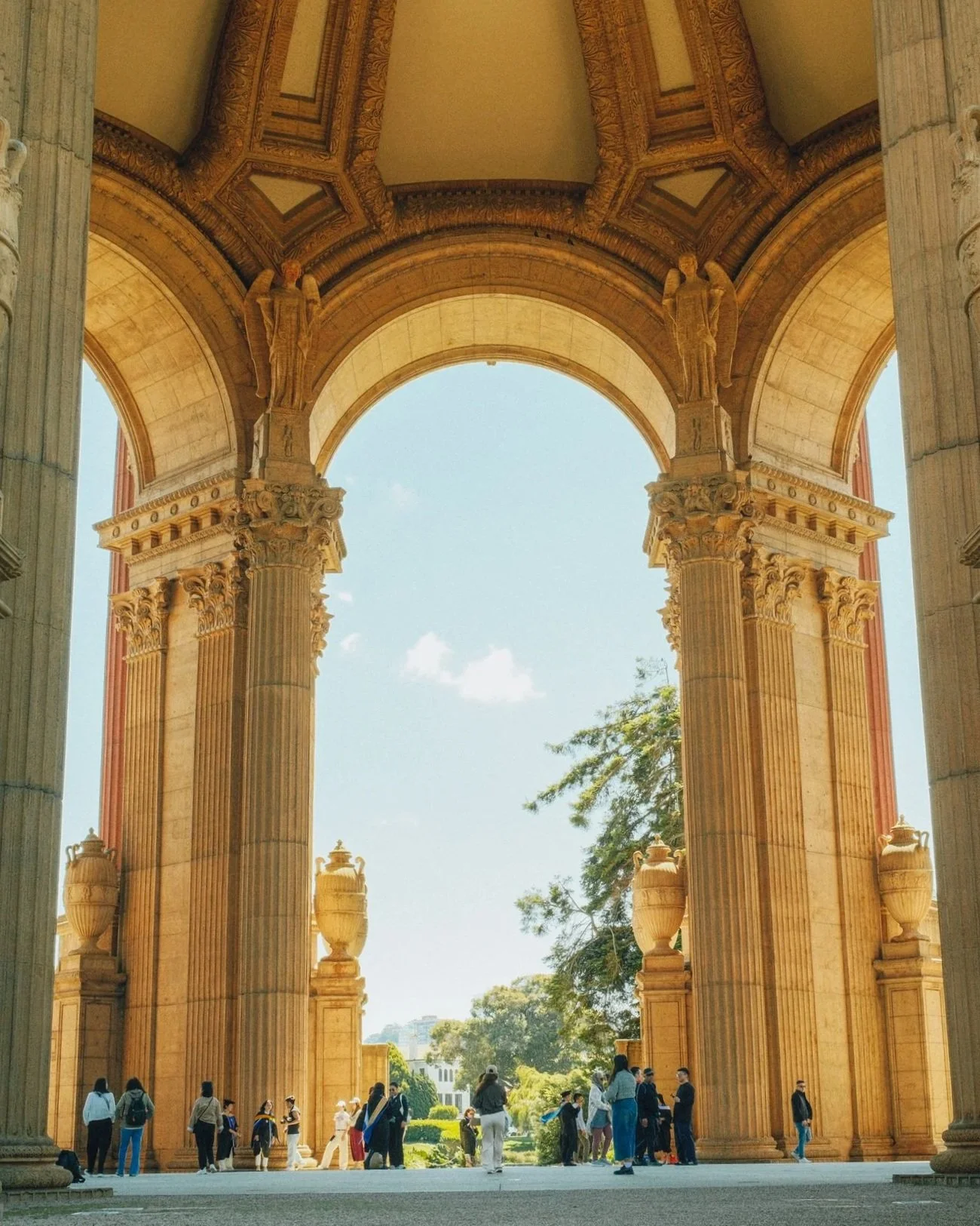 People walking under large, ornate stone archway with two towering columns decorated with intricate carvings and large urns, with a clear blue sky and trees in the background.