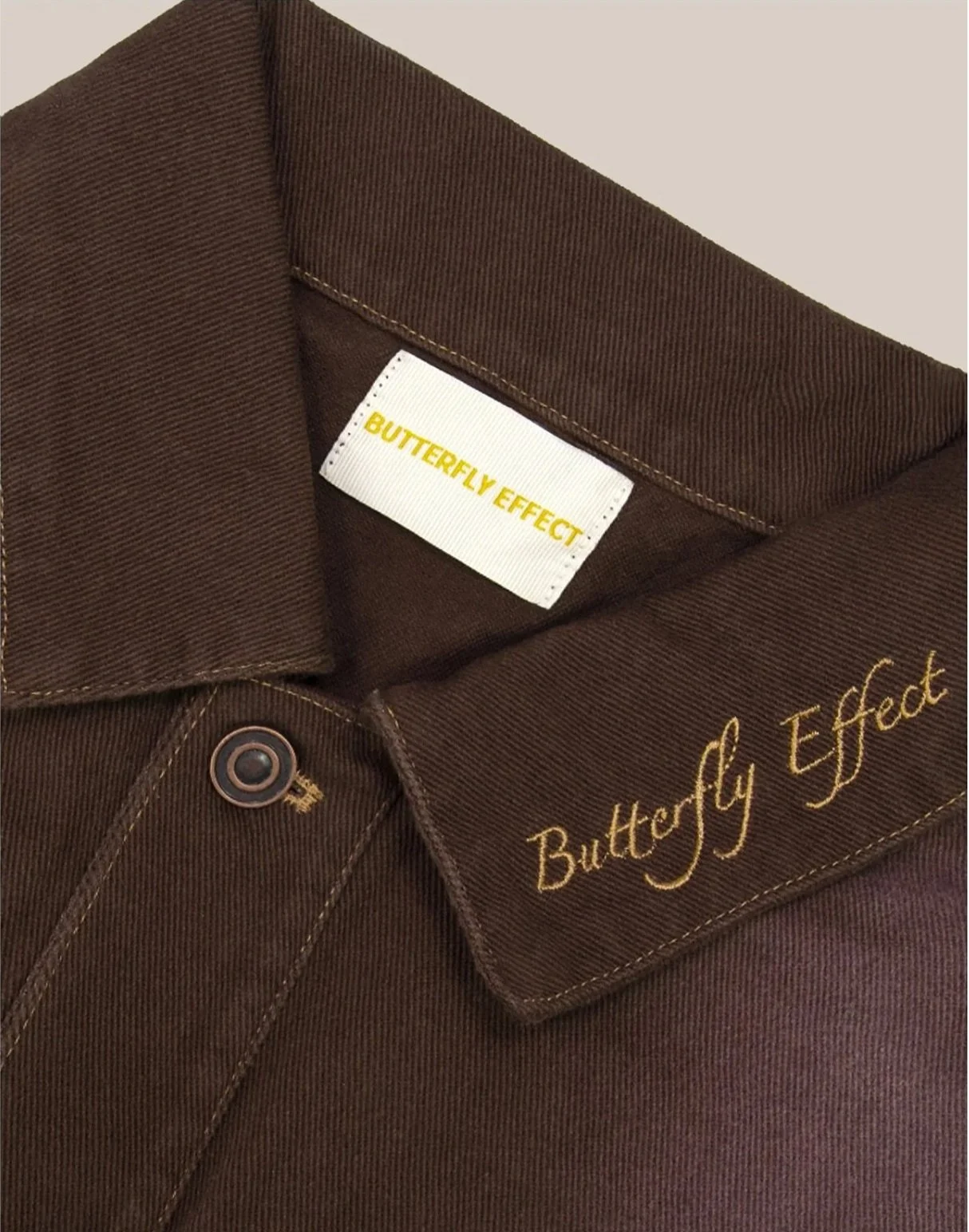 Close-up of a brown shirt collar with a white label that says "Butterfly Effect" in yellow text and embroidery on the pocket that reads "Butterfly Effect" in yellow cursive.