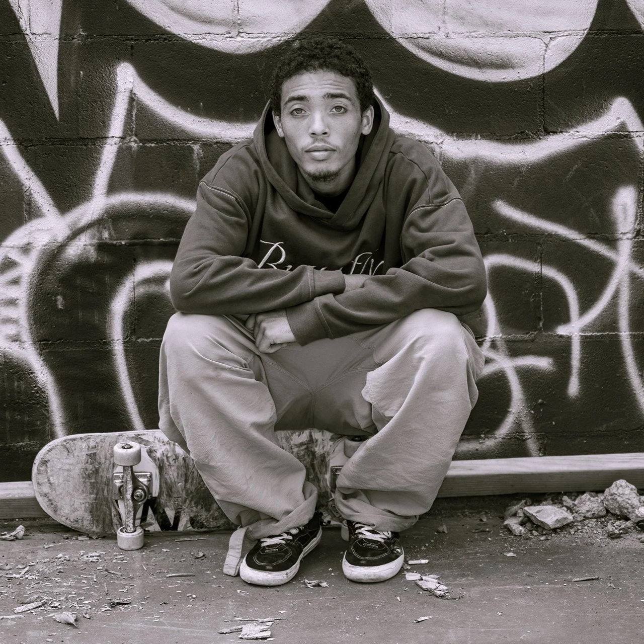 A young man sitting on the ground with a skateboard beside him in front of a painted brick wall.