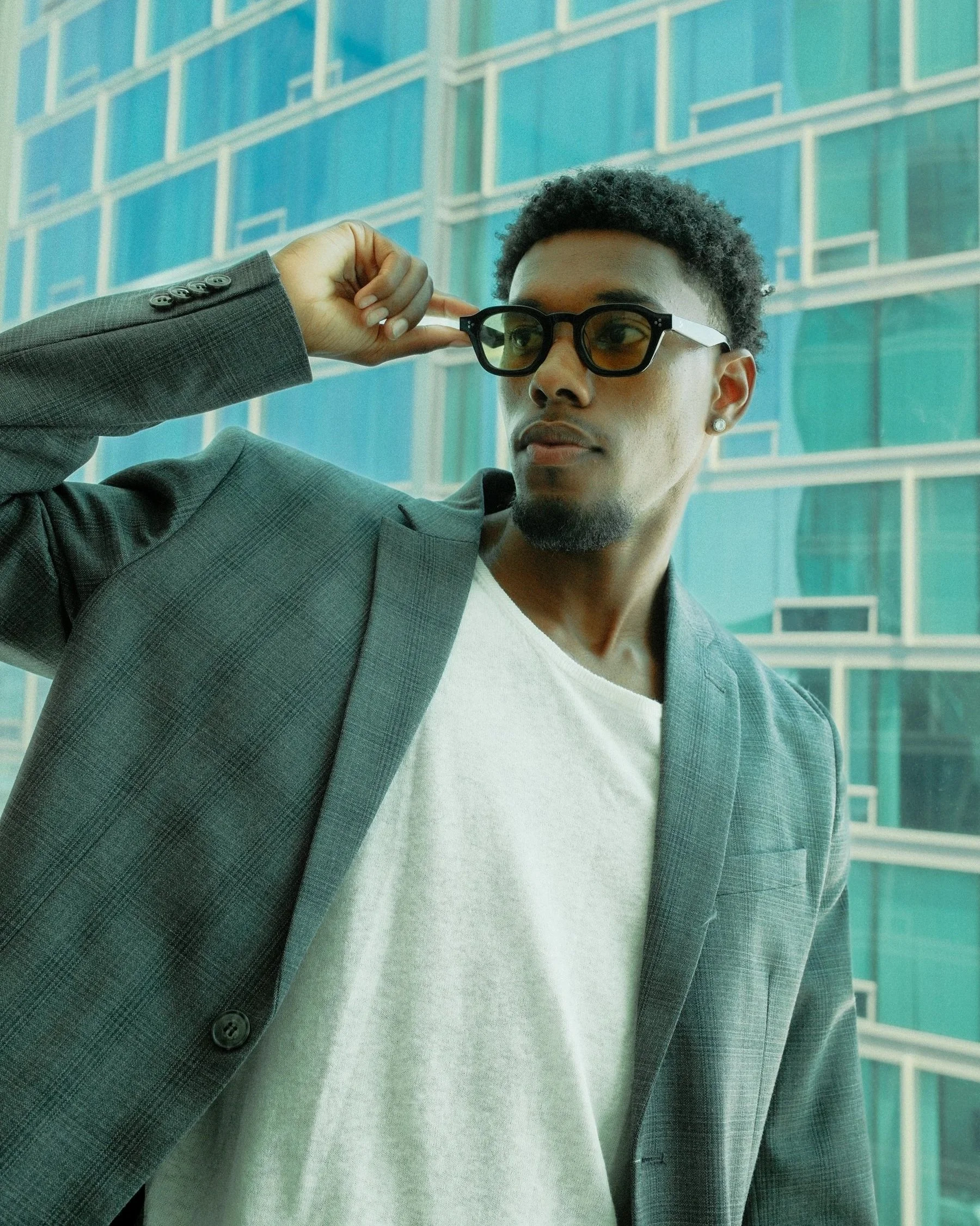 A young man wearing a gray checkered blazer, white shirt, and sunglasses, standing outdoors in front of a modern glass building.