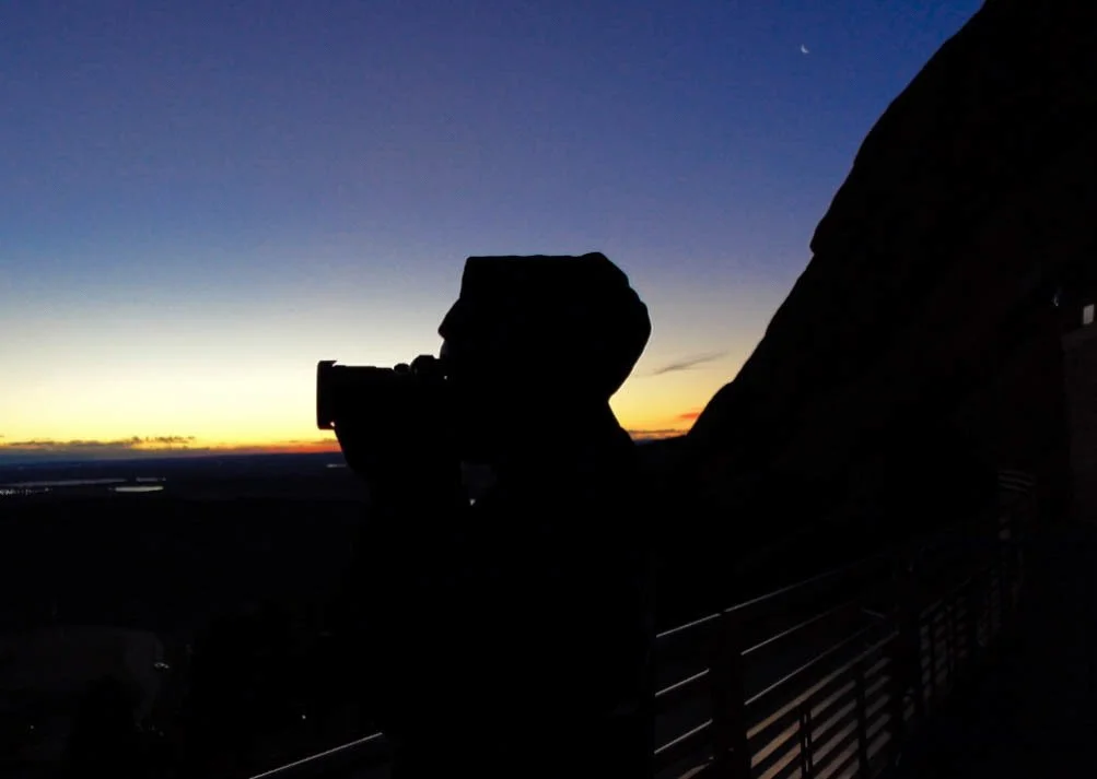 Silhouette of a person taking a photo with a camera on a balcony during sunset, with a mountain or large rock formation to the right and a clear sky transitioning from orange to dark blue.