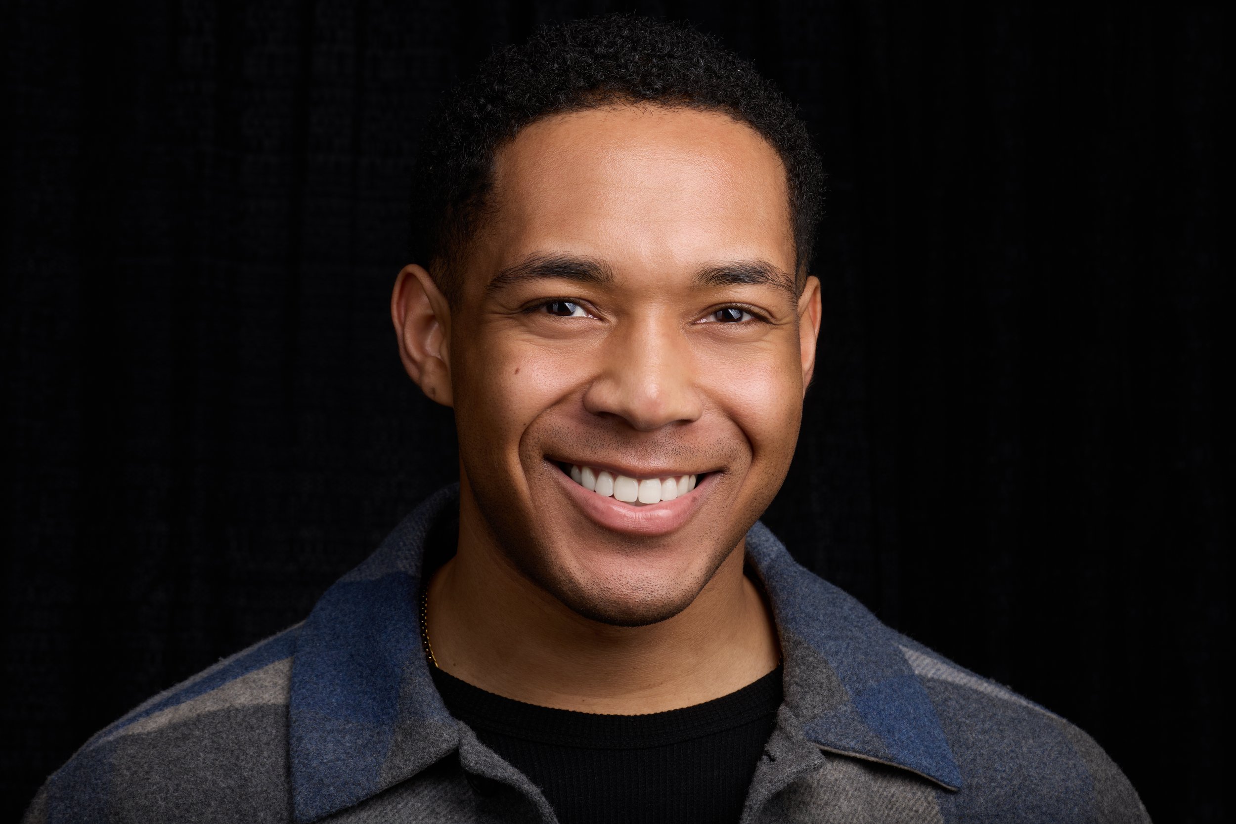 Close-up portrait of Joshua Bradley with short curly hair, wearing a dark t-shirt and a blazer, against a black background.