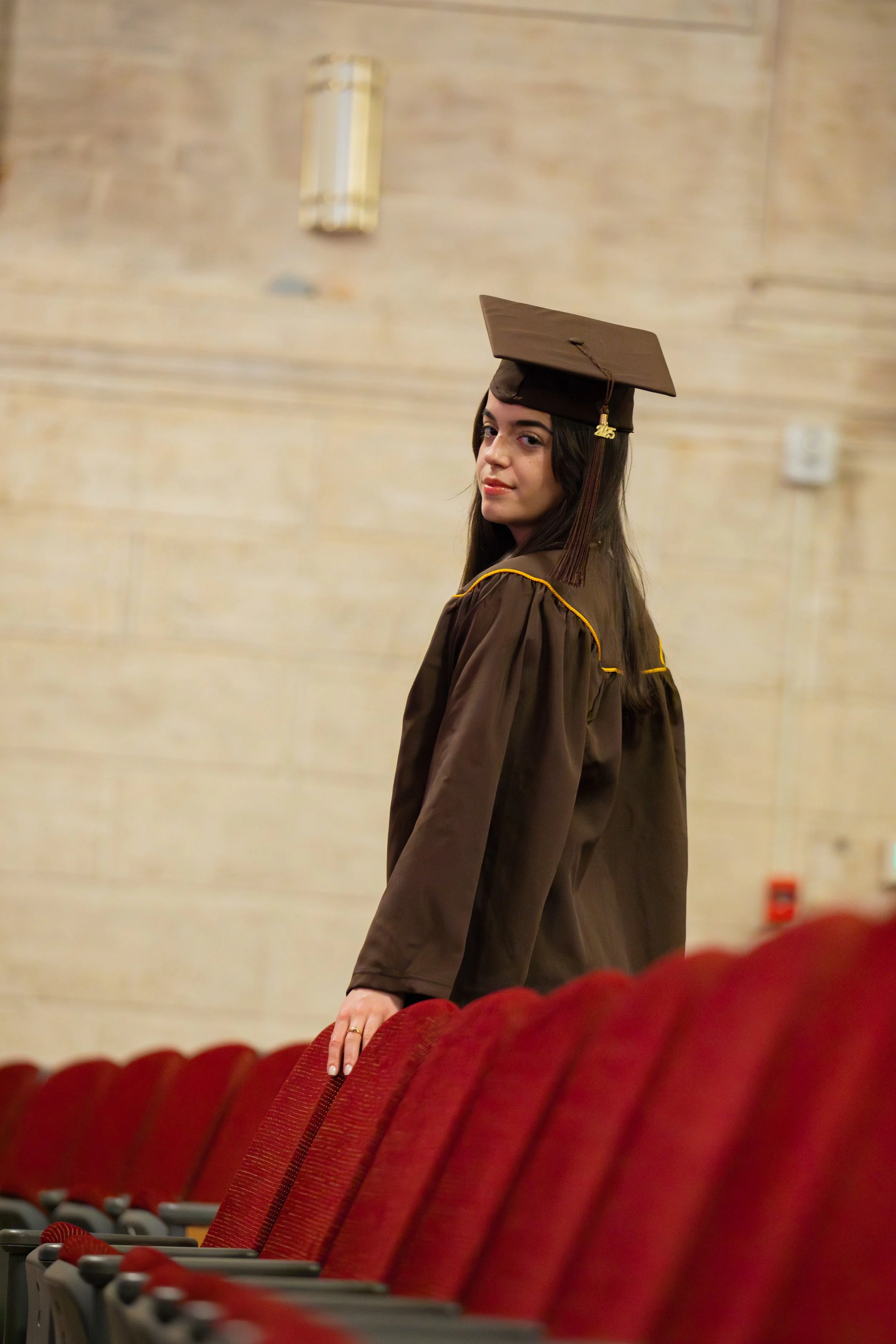 A young woman in a graduation cap and gown standing in an auditorium. She is looking over her shoulder at the camera with a slight smile, with red theater seats in the foreground.