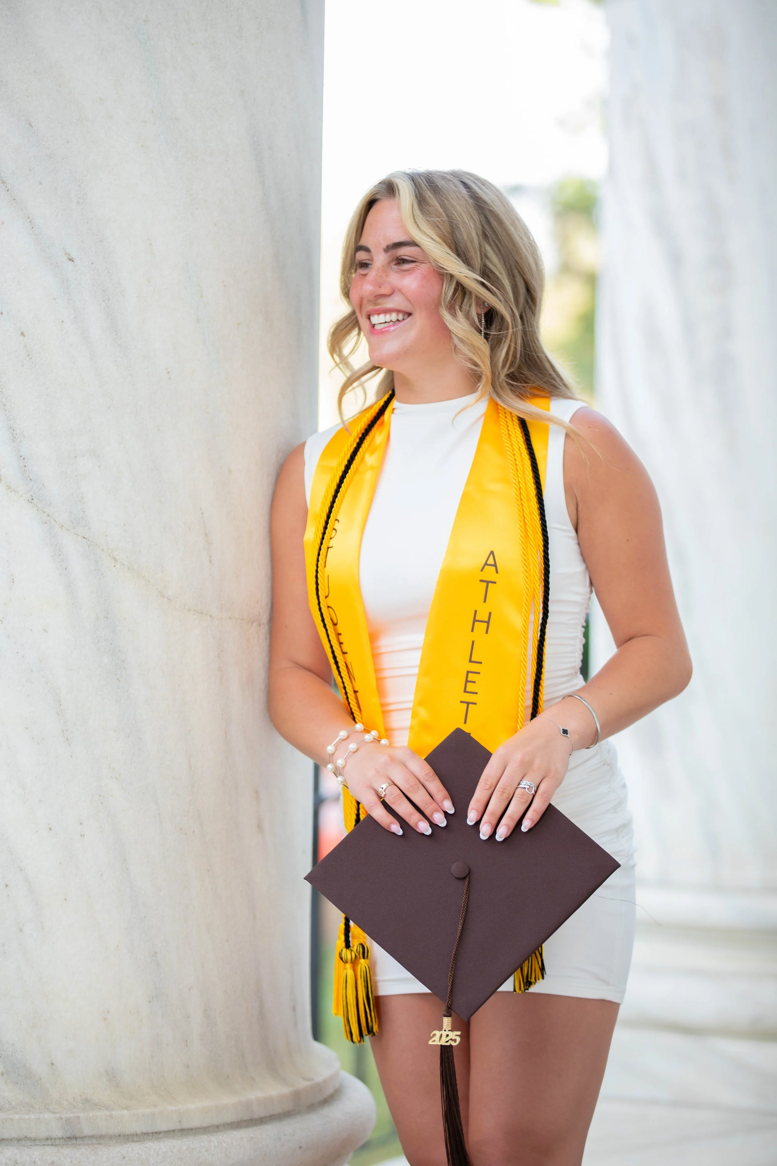 A young woman with blonde hair smiling, wearing a white dress, and a yellow honor cord with black accents. She holds a black graduation cap with a 2023 charm, standing outdoors near a large stone column.