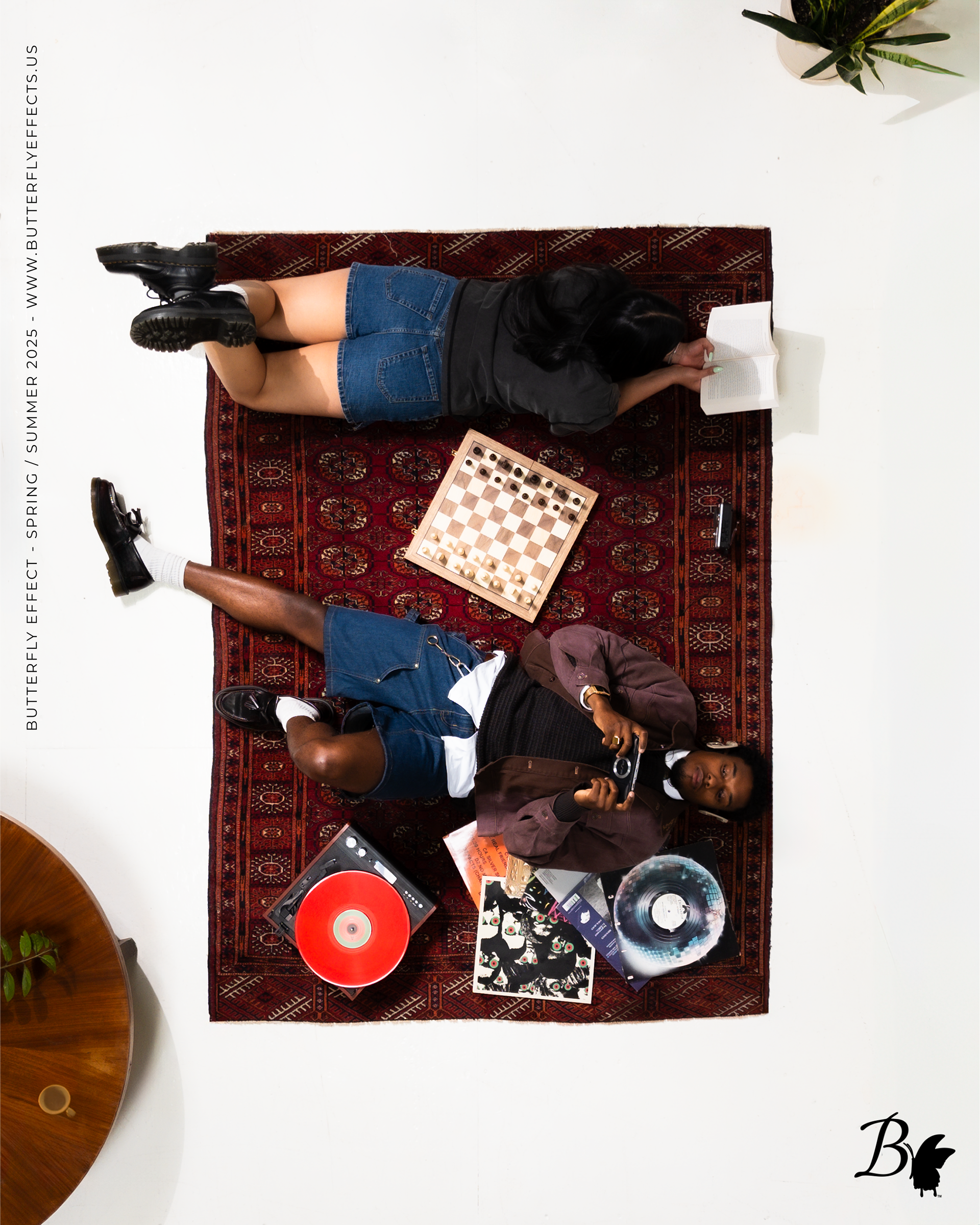Top-down view of a person lying on a red patterned rug on a white floor, reading a book. They are wearing black shorts, a black t-shirt, and have long black hair. Surrounding items include a potted plant in the top right corner, a vinyl record and ma