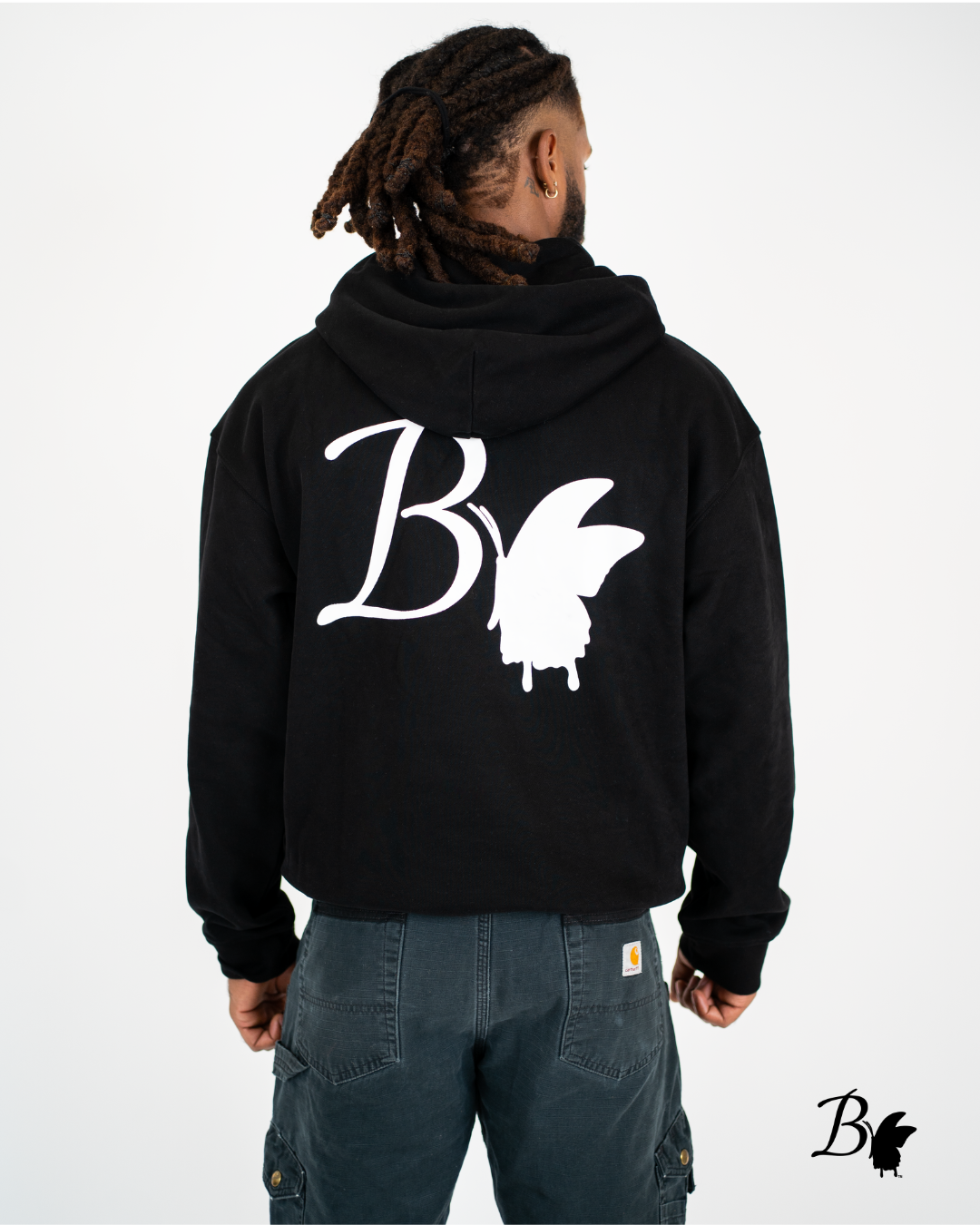 A man with dreadlocks wearing a black hoodie with a large white 'B' and a butterfly graphic on the back, standing with his back to the camera against a white background.