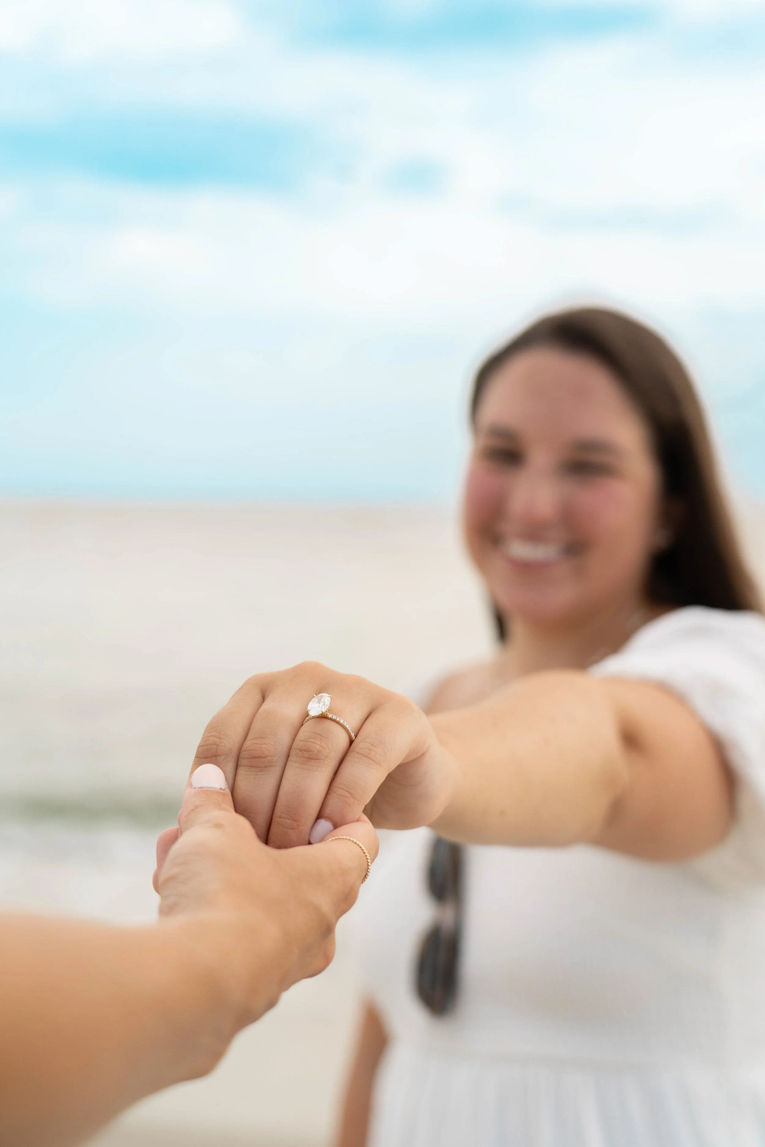 A woman smiling and showing her engagement ring as she holds hands with another person on a beach with a cloudy sky in the background.