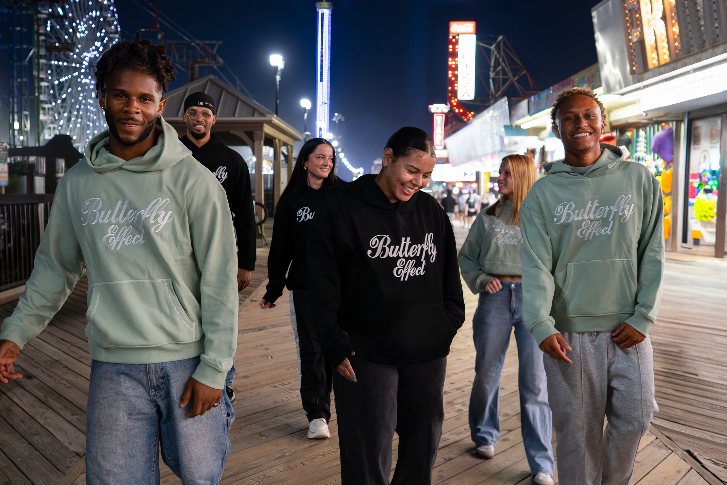 Group of five young adults walking at night at an amusement park or fairground. They are smiling and wearing casual clothing, some with hoodies displaying the words "Butterfly Effect." Bright lights and rides are visible in the background.