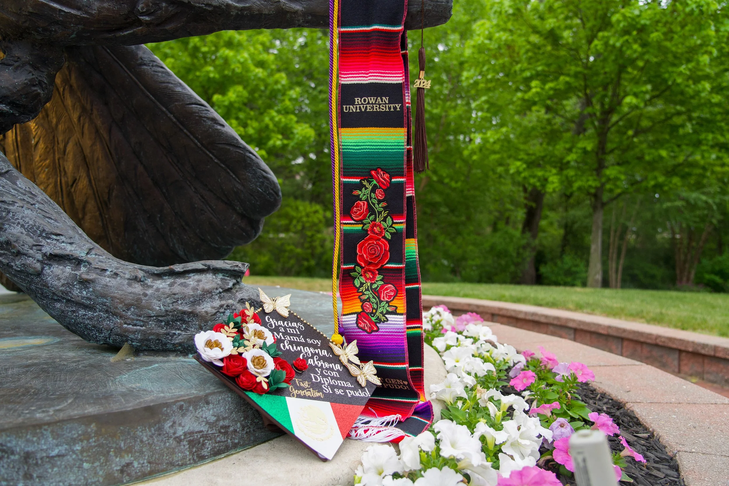 A memorial with a bronze statue of a hand holding a flower, surrounded by colorful flowers and decorative items. Hanging from the hand is a traditional Mexican woven sash with the words "ROWAN UNIVERSITY" and an embroidered floral design. Various flo