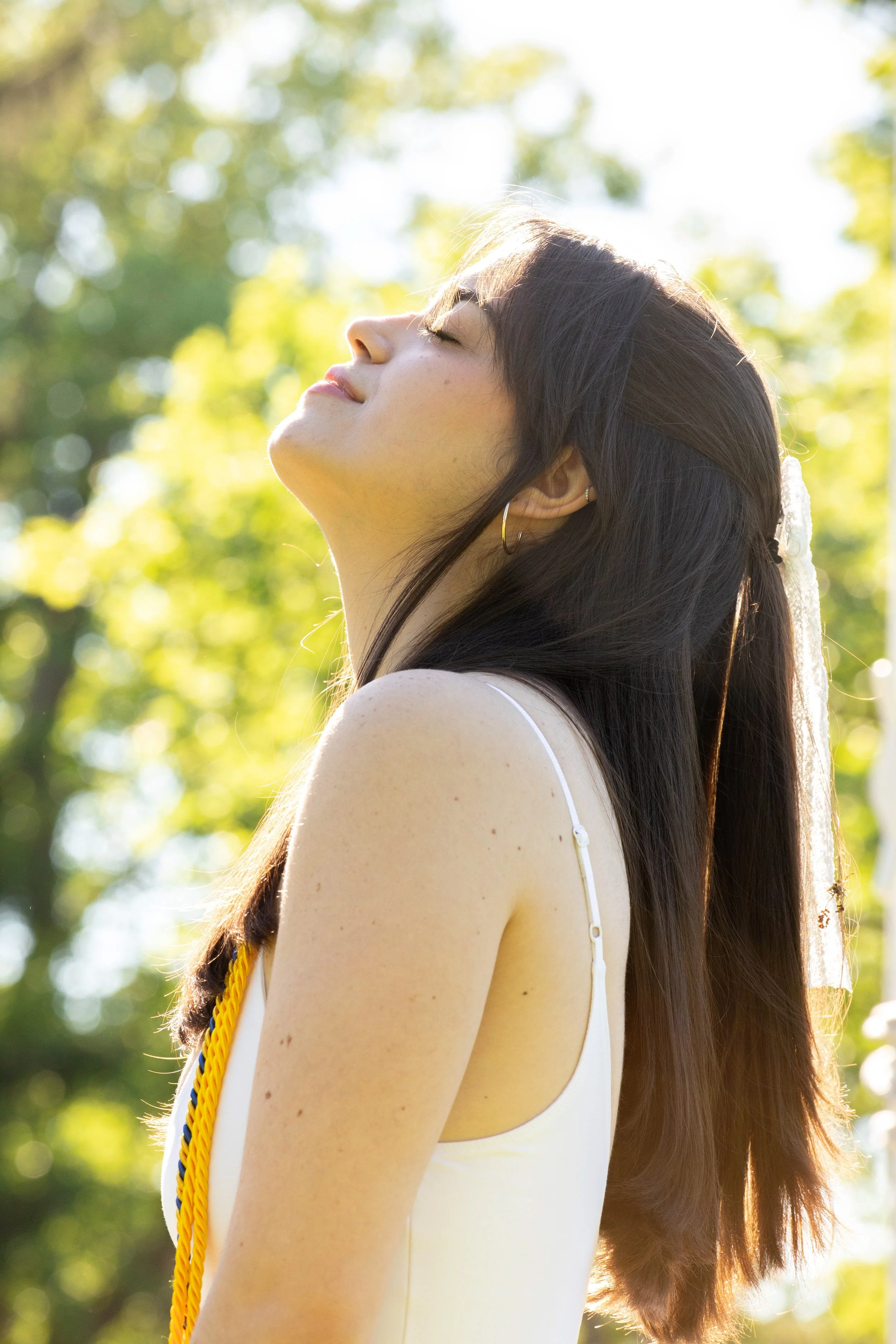 A young woman with long brown hair, wearing a white dress and gold hoop earrings, standing outdoors with her eyes closed and facing sunlight, in a park with green trees.