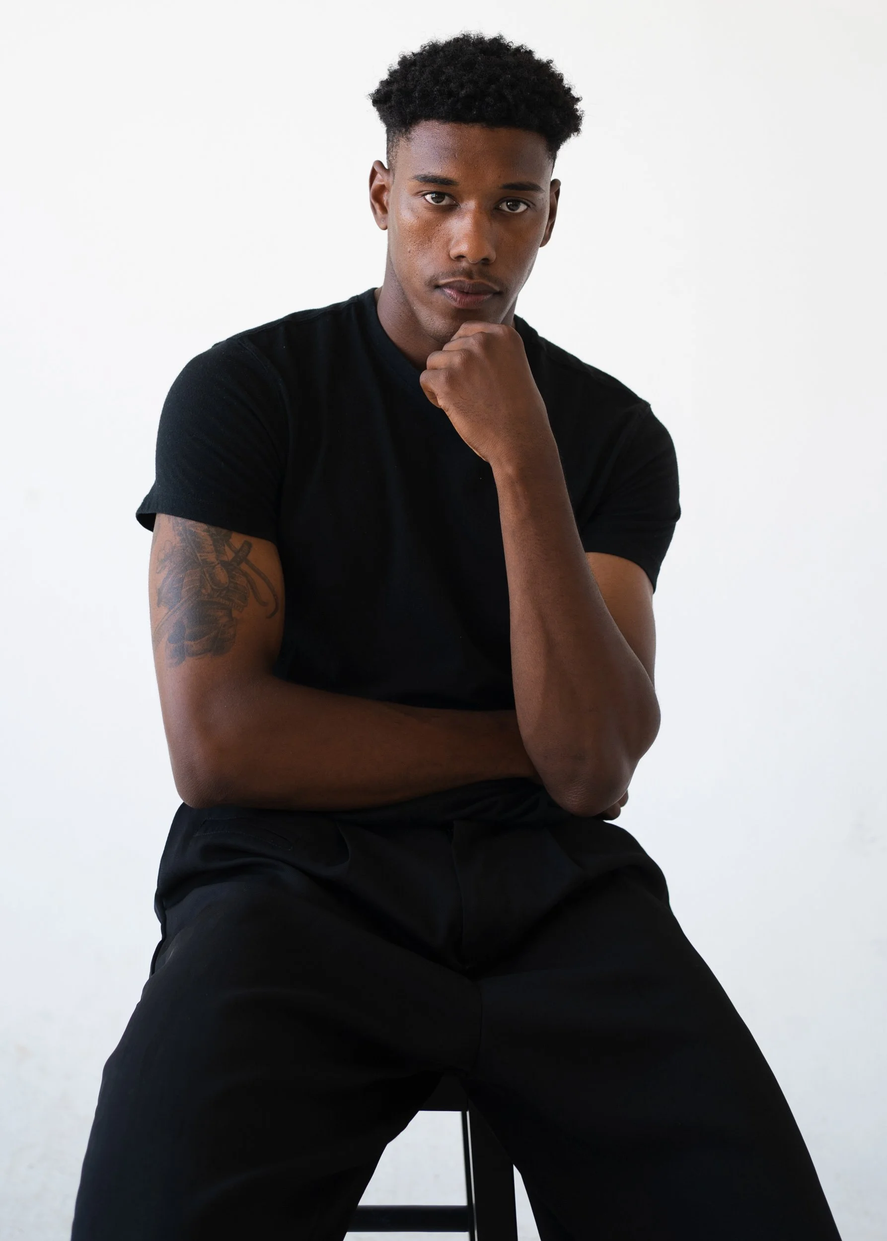 Young man with short curly hair, wearing a black t-shirt, sitting on a black stool against a white background, with his right hand under his chin and left arm crossed over his chest, looking at the camera.