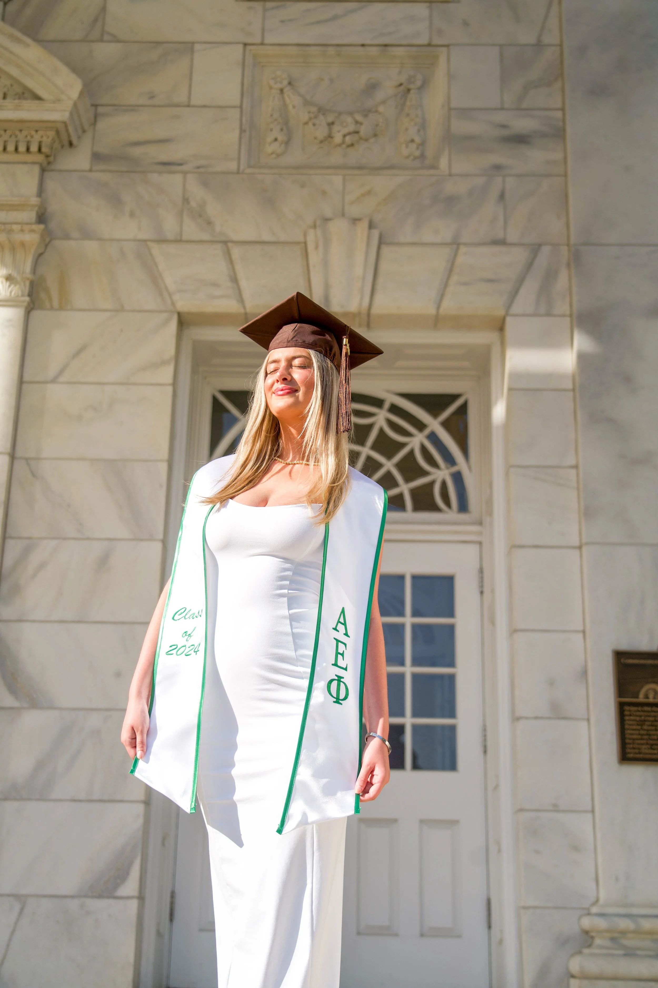 A young woman in a white dress and graduation cap stands in front of a historic building, smiling with her eyes closed. She is wearing a sash that reads 'Class of 2024' and features the Greek letters Phi Sigma Phi.