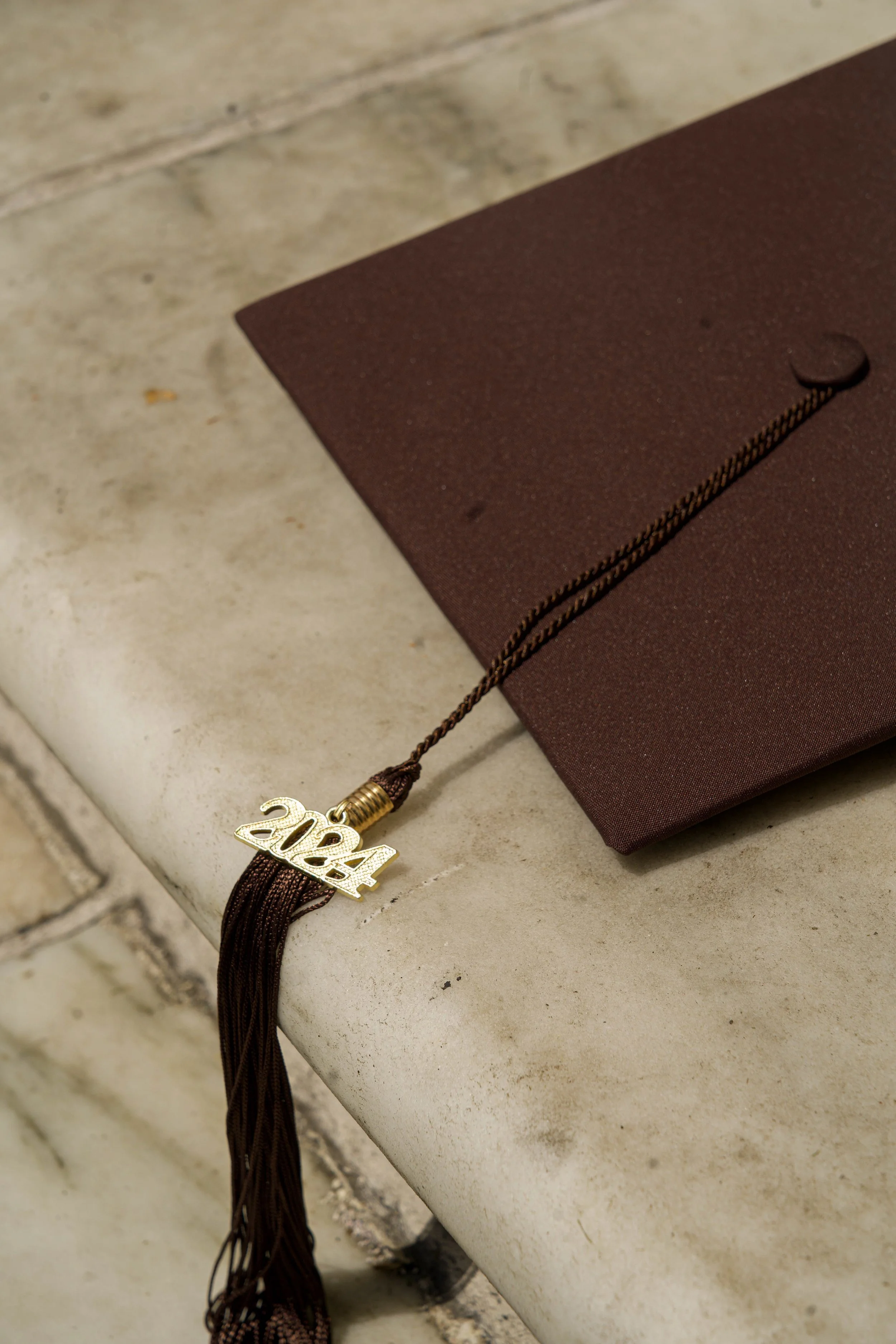 A brown graduation tassel with a gold charm displaying the year 2024, resting on a brick surface with a maroon object nearby.