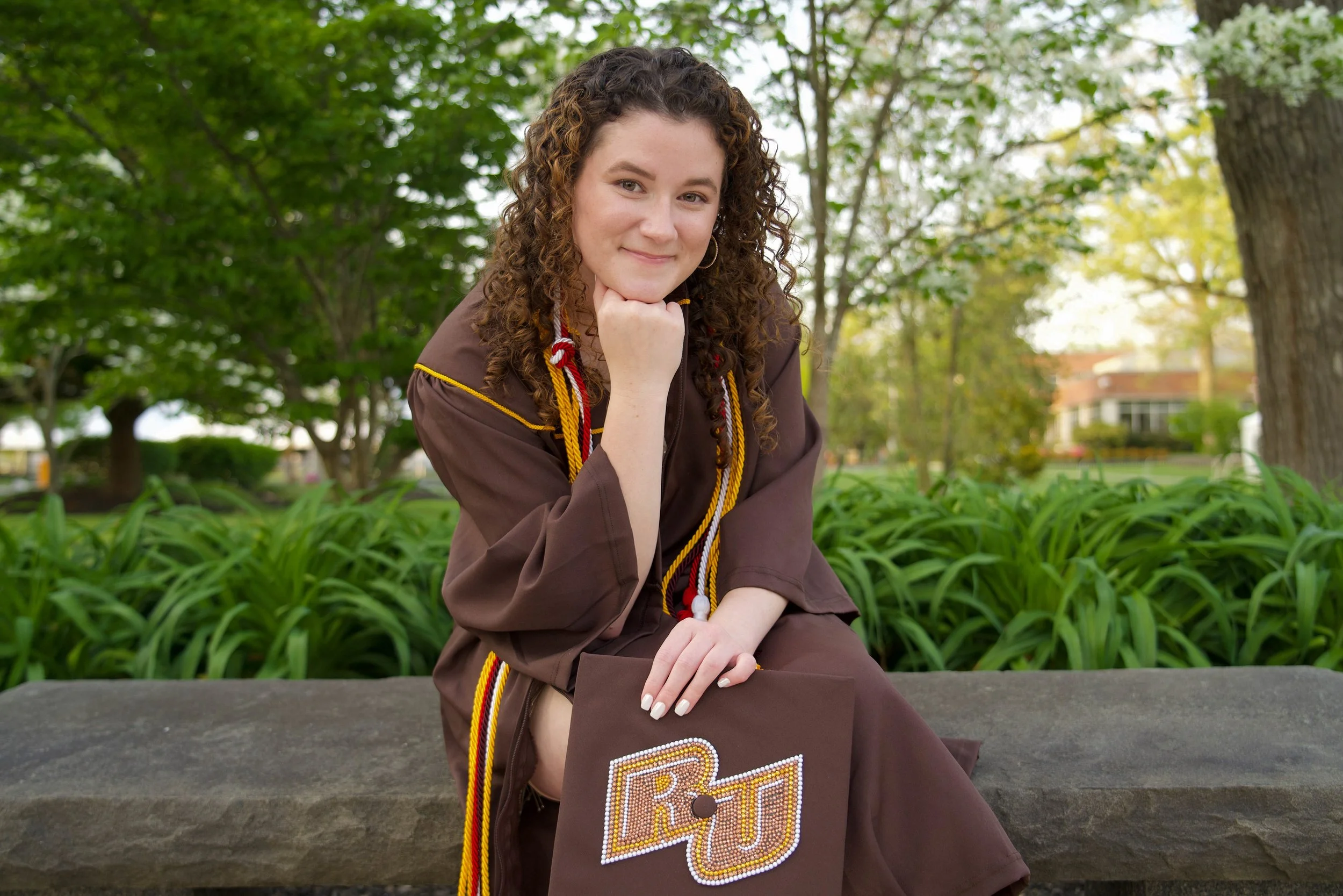 A young woman with curly brown hair, wearing a brown graduation gown and honor cords, sitting outdoors on a park bench with green trees and a building in the background.