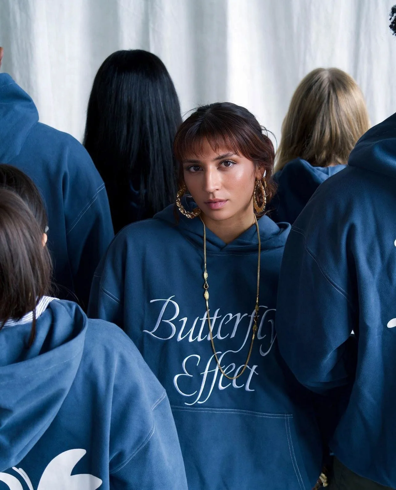 Young woman with brown hair, wearing hoop earrings, gold necklace, and a blue hoodie with white cursive text, standing among other people in similar hoodies.
