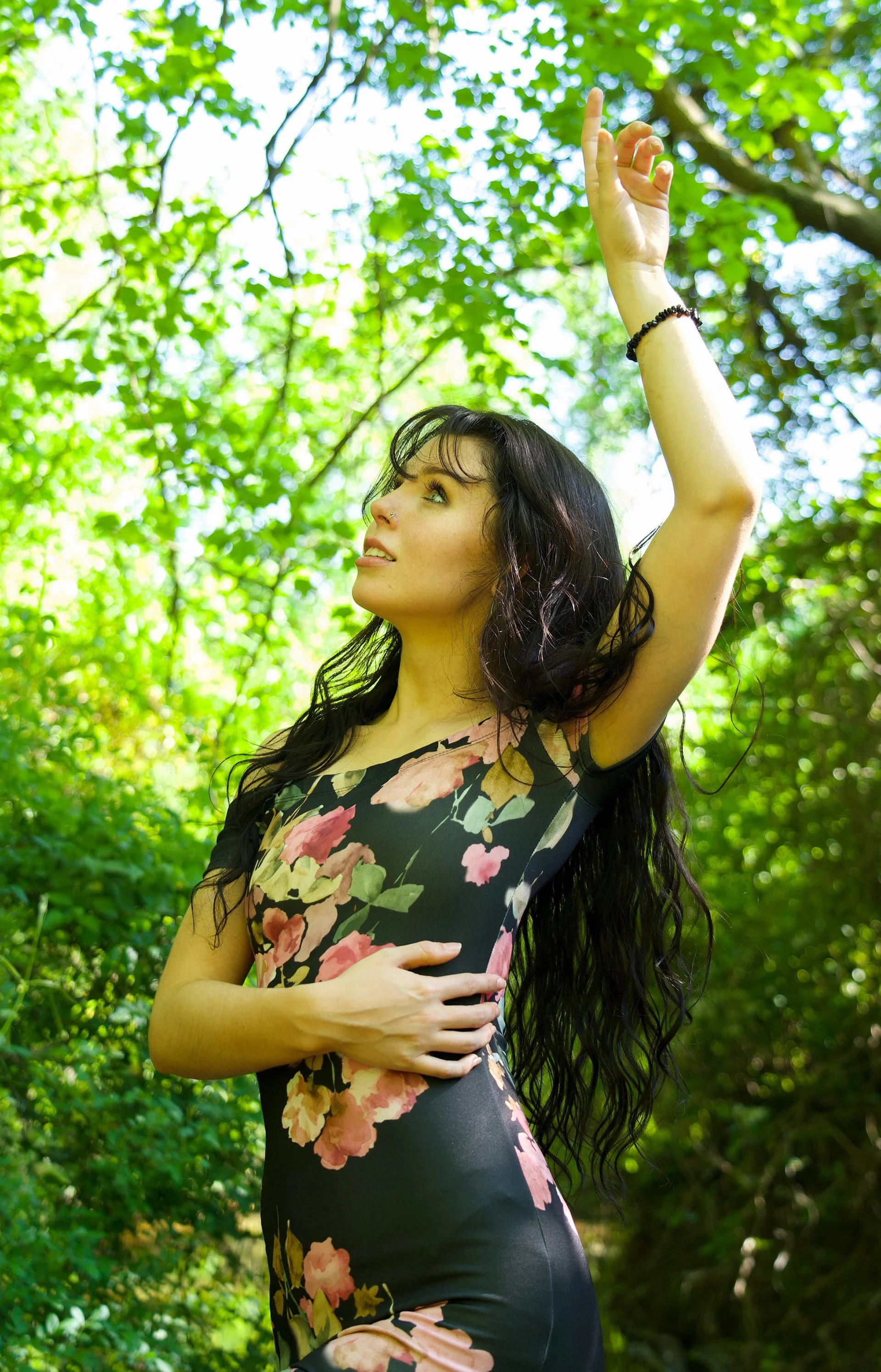 A woman with long dark hair in a floral dress standing outdoors among green trees, raising her right hand with an open palm and index finger pointing upward, looking upward with a serene expression.